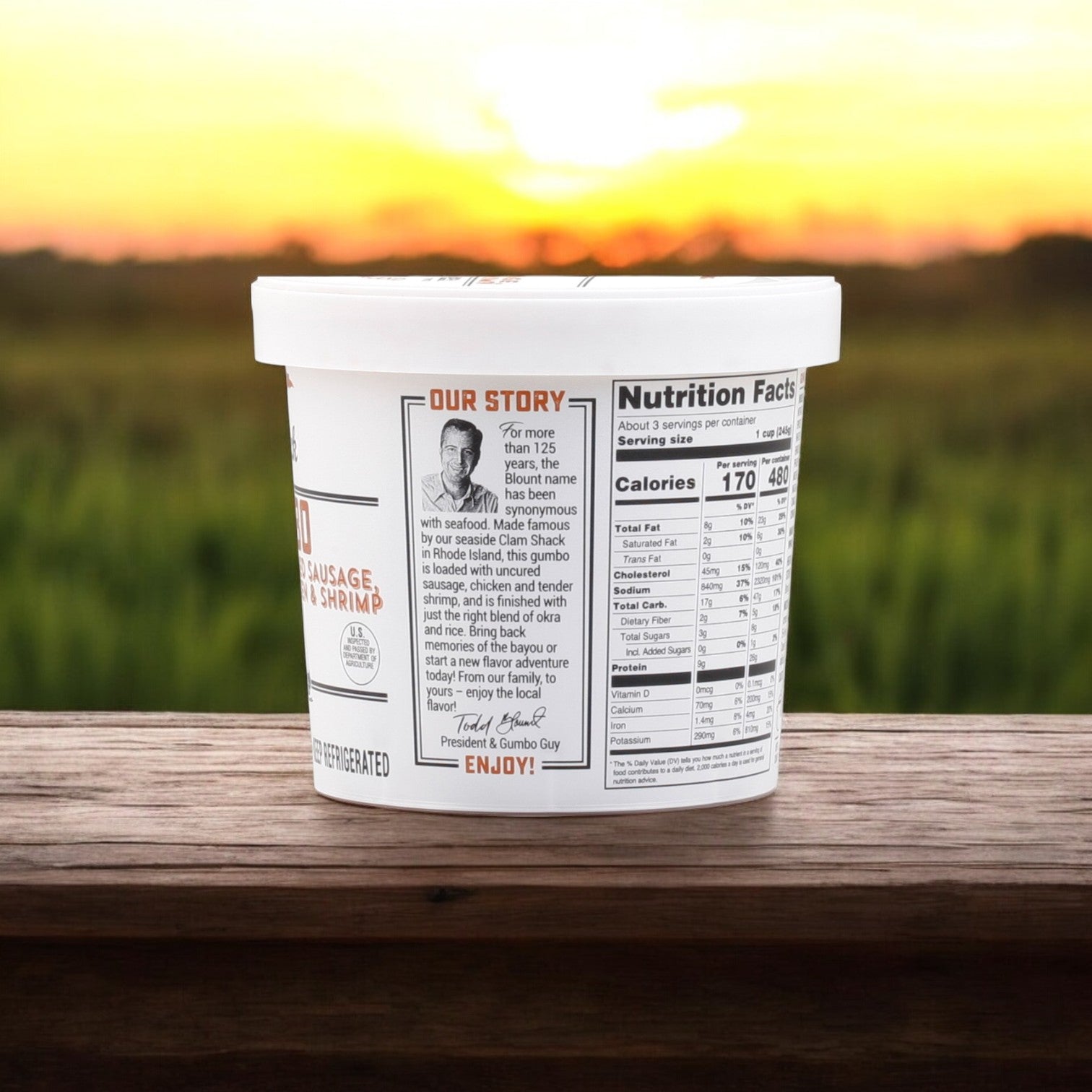 A cup of Blount Clam Shack Gumbo (Uncured Sausage, White Meat Chicken & Shrimp), with nutrition info and a Creole spices story, sits on a wooden surface against a blurred outdoor backdrop at sunset.