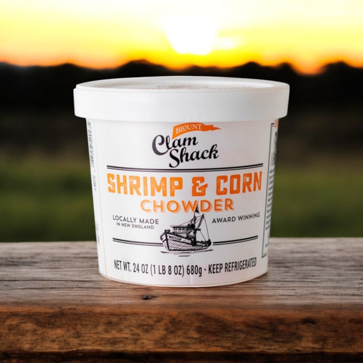 A 24 oz. tub of Blount Clam Shack Shrimp & Roasted Corn Chowder by Blount Fine Foods, a delightful seafood treat, rests on a wooden surface with a sunset backdrop.