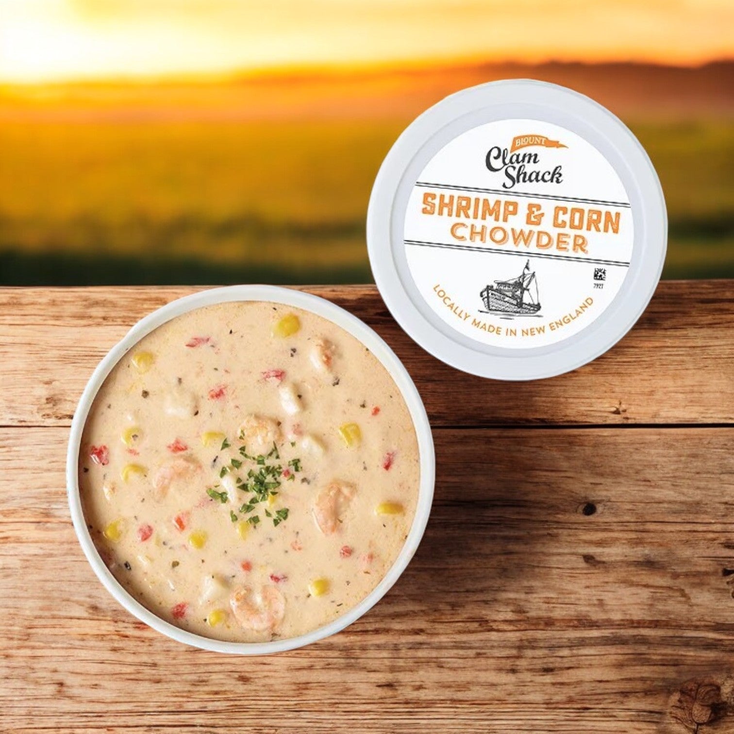 A 24 oz. bowl of Blount Clam Shack Shrimp & Roasted Corn Chowder from Blount Fine Foods rests on a wooden table, its aromatic richness blending with the sunset's golden hues—a genuine seafood delight in every spoonful.