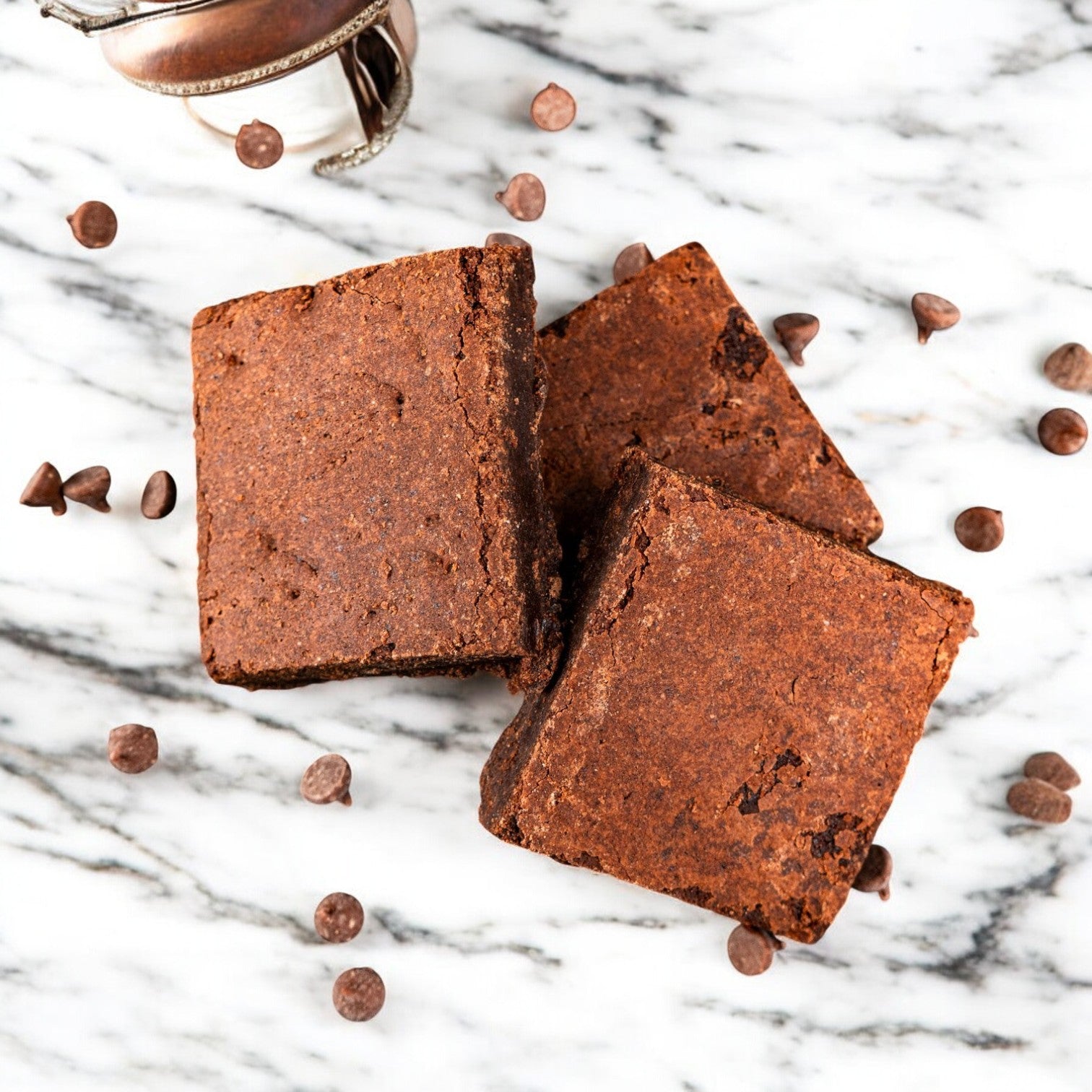 Three fudgy brownies from OMG It's Gluten Free's Brownie Fudge pack lie on a marble surface, surrounded by rich chocolate chips.