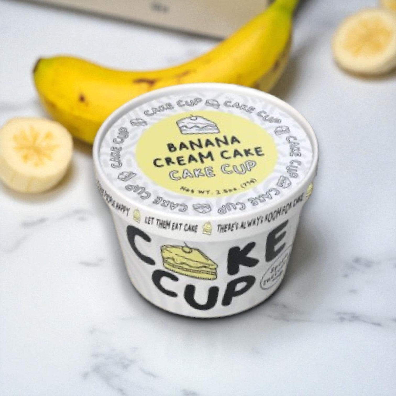 The Eat Cup Cake "Banana Cream Cake – 2.5 oz (Spoon Included)" is displayed on a marble surface, accompanied by a whole banana and a banana slice, highlighting this ready-to-eat dessert in its individual cup.