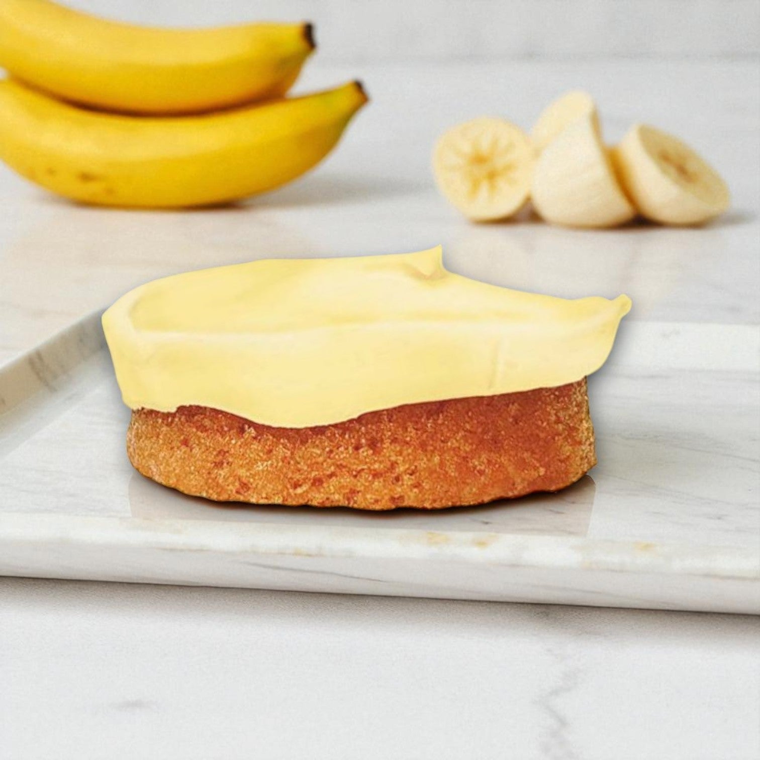 The Eat Cup Cake Banana Cream Cake – 2.5 oz (Spoon Included) features yellow frosting and comes ready to eat, making it a tempting dessert, with sliced and whole bananas in the background on a marble surface.