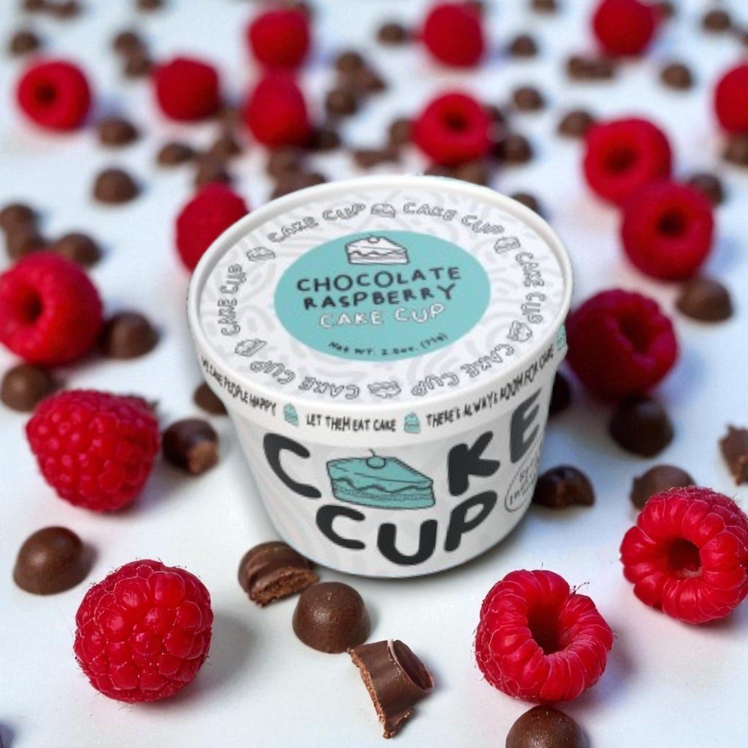 A 2.5 oz Eat Cup Cake Chocolate Cake Cup (spoon included) by Eat Cup Cake is displayed on a white surface with fresh raspberries and chocolate pieces, making it a perfect single-serve dessert.
