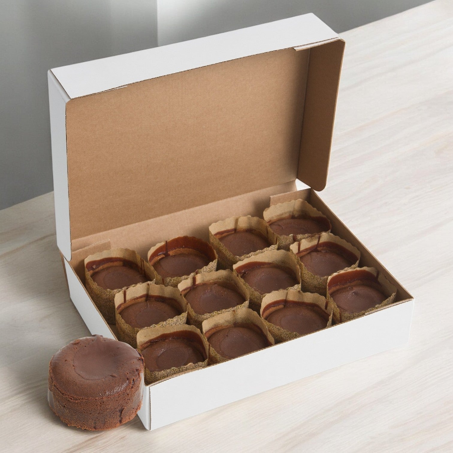 A white box labeled "Sysco Cake Fudge Divine Lava 3", 5 oz. - 1 Case, 24 Fudge Cakes" on a wooden surface holds nine round chocolate desserts with molten fudge centers, accompanied by two more treats beside it.