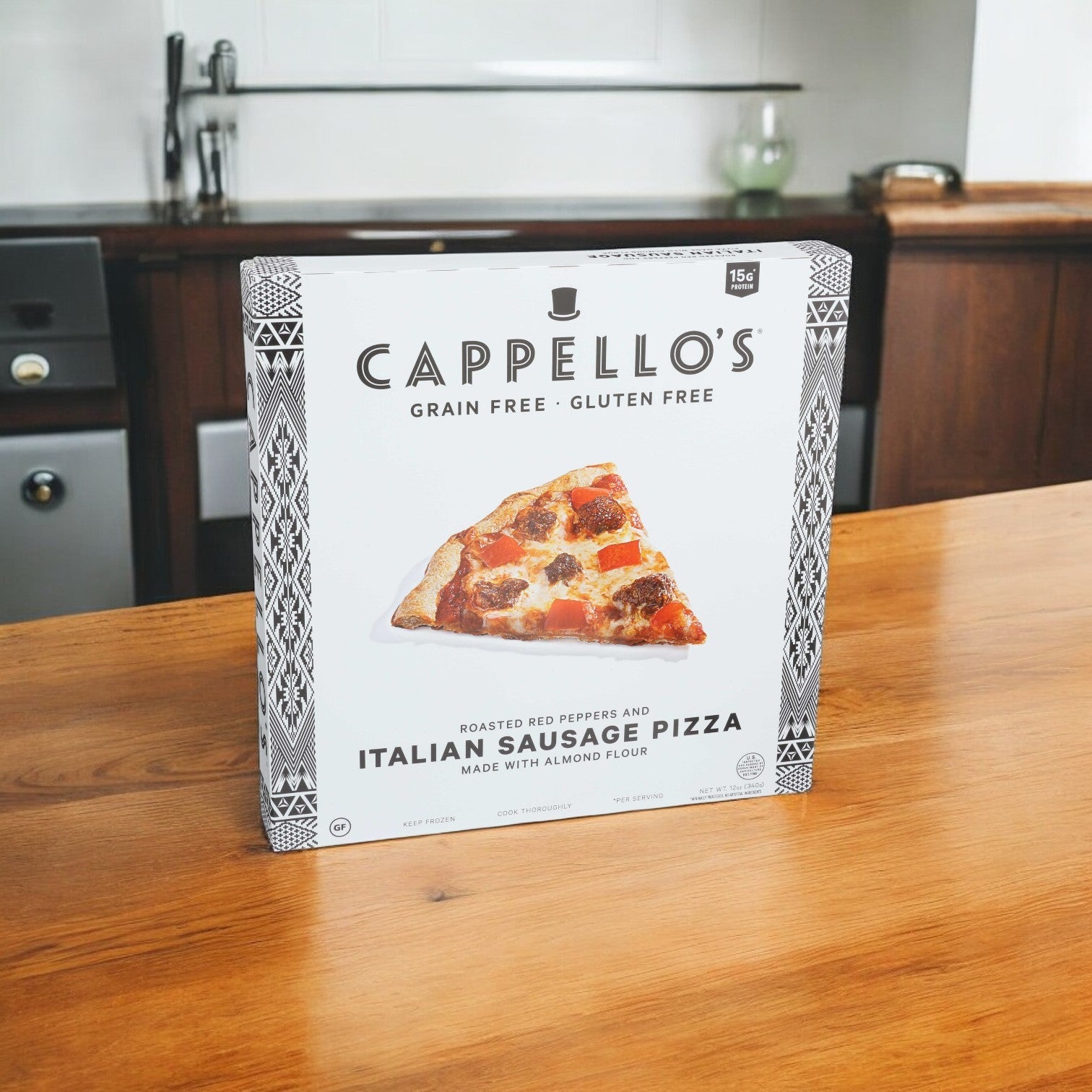 A box of Cappello's Italian Sausage with Roasted Red Peppers Pizza, featuring an almond flour crust, rests on a wooden table set against a kitchen background.