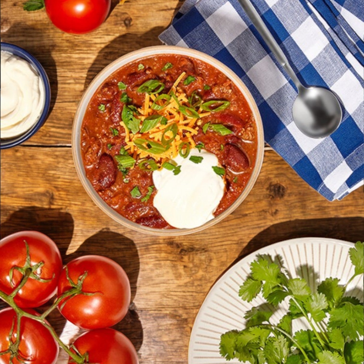 A bowl of Bistro Soups by Vienna Beef – Cheli’s Chili sits on a wooden table, topped with cheese, sour cream, and herbs. Nearby are juicy tomatoes, fresh cilantro, a napkin with a spoon, and an extra bowl of sour cream for that Chicago-style flavor twist.