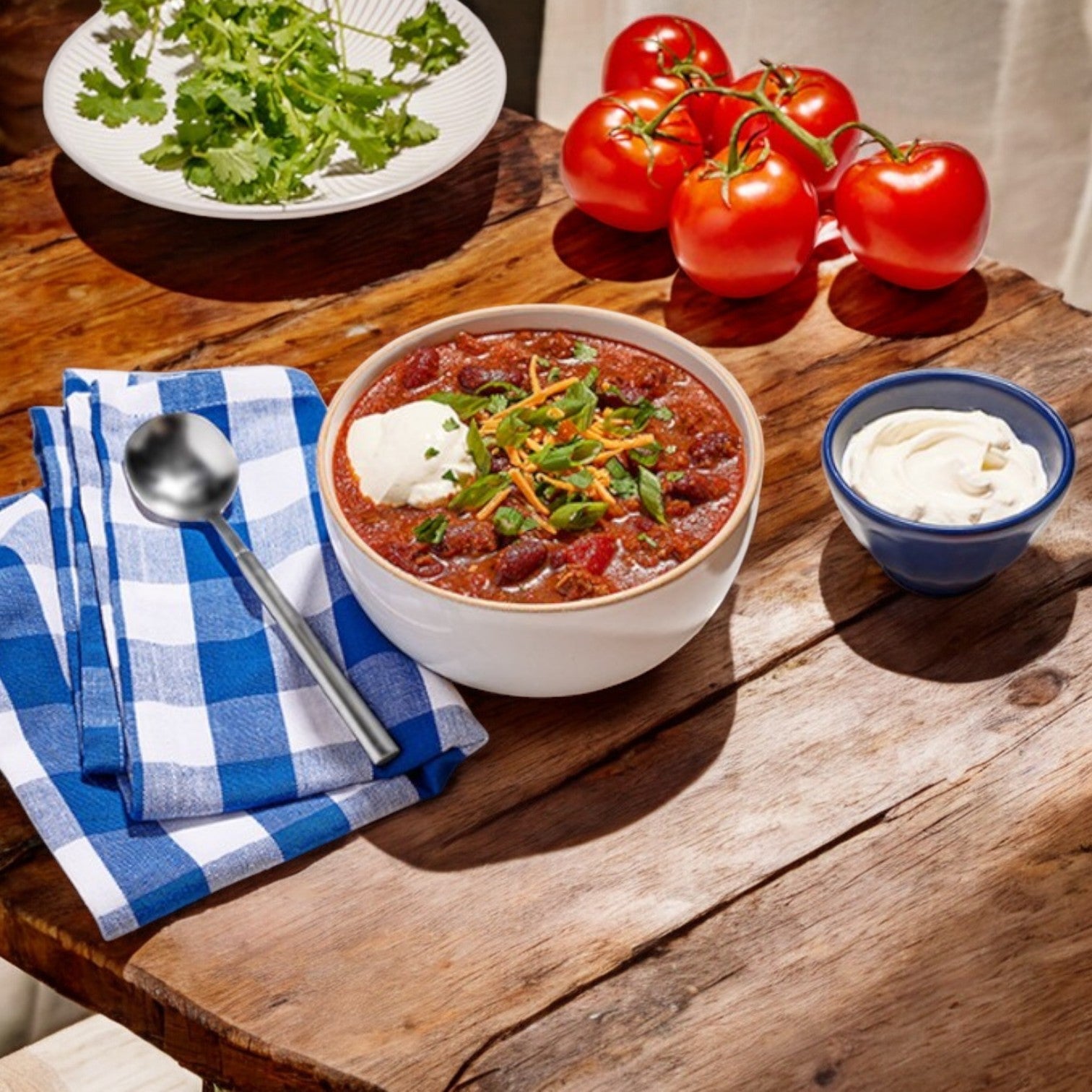 A bowl of Cheli’s Chili from Vienna Beef's Bistro Soups sits on a wooden table, garnished with sour cream and green onions, surrounded by tomatoes, cilantro, a blue bowl of sour cream, a spoon, and a blue checkered napkin. Its Chicago-style flavor adds zest to every bite.