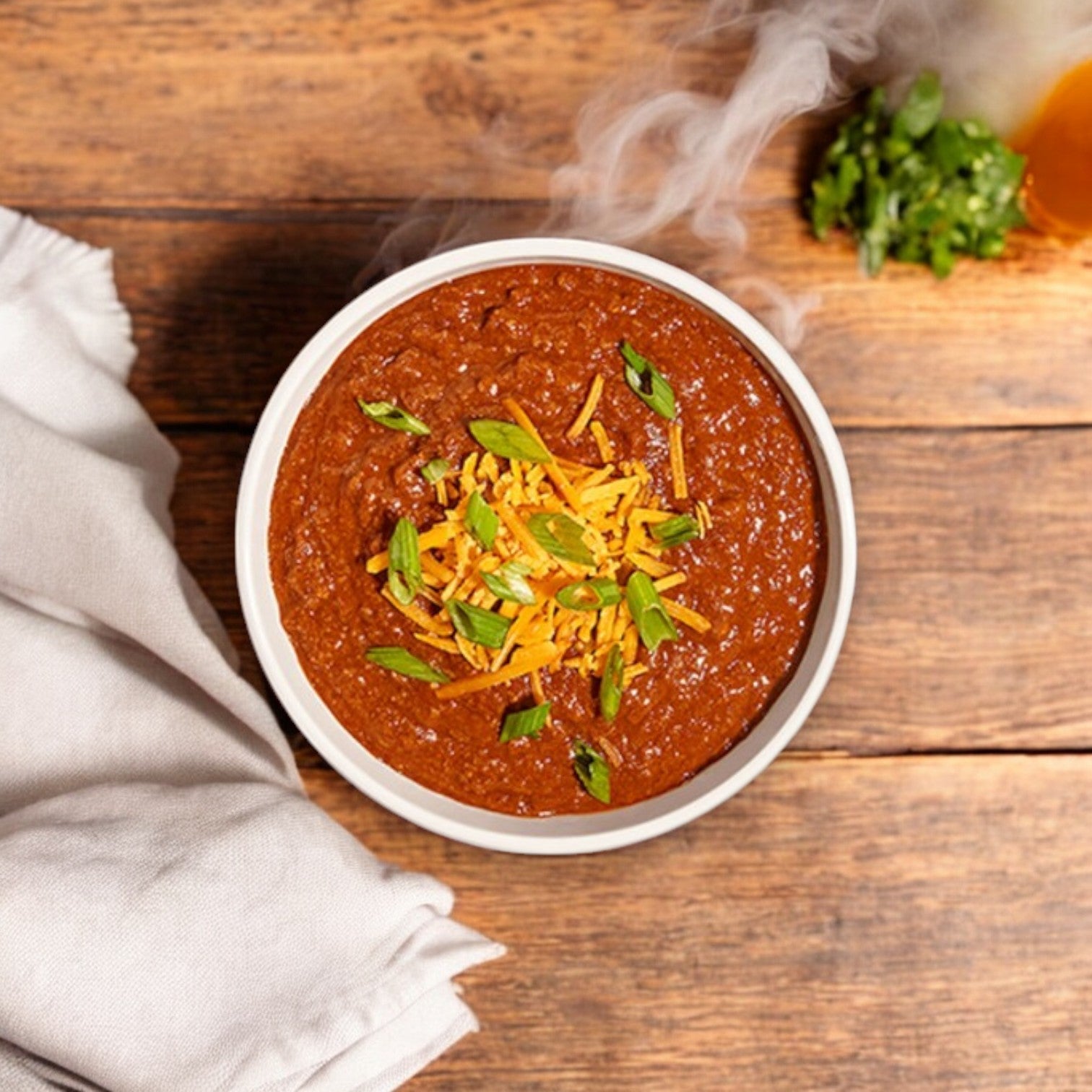 A steaming bowl of Bistro Soups by Vienna Beef - Chili Dog Chili, garnished with shredded cheese and green onions, sits on a wooden table next to a folded white napkin. Chopped herbs and a slice of orange offer a delightful mix of flavors and colors.