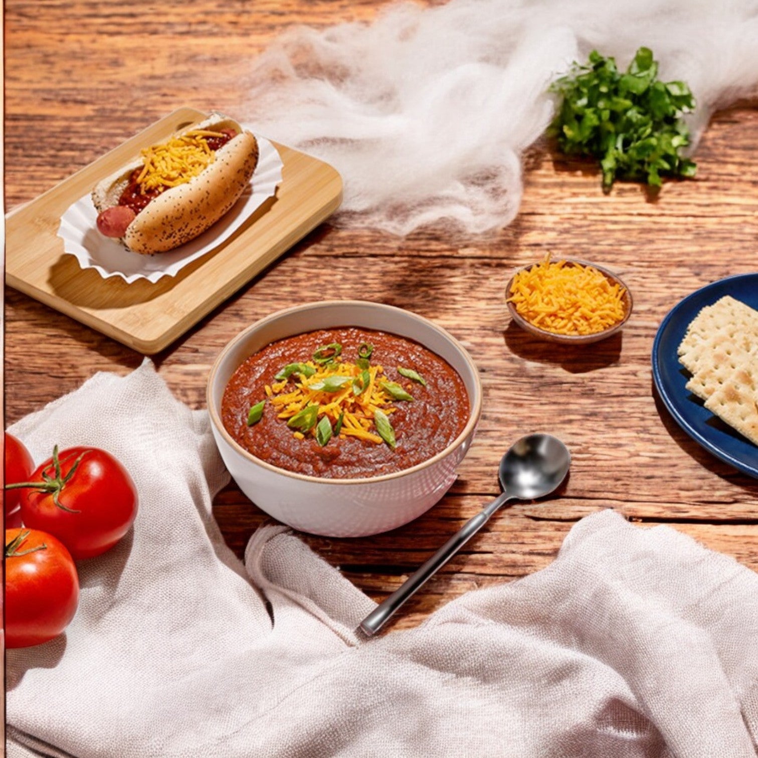 A bowl of Vienna Beef Bistro Soups Chili Dog Chili is topped with cheese and green onions on a wooden table. Nearby, a Vienna Beef hot dog with cheese, tomatoes, crackers, and extra cheese awaits. A spoon rests beside the chili.