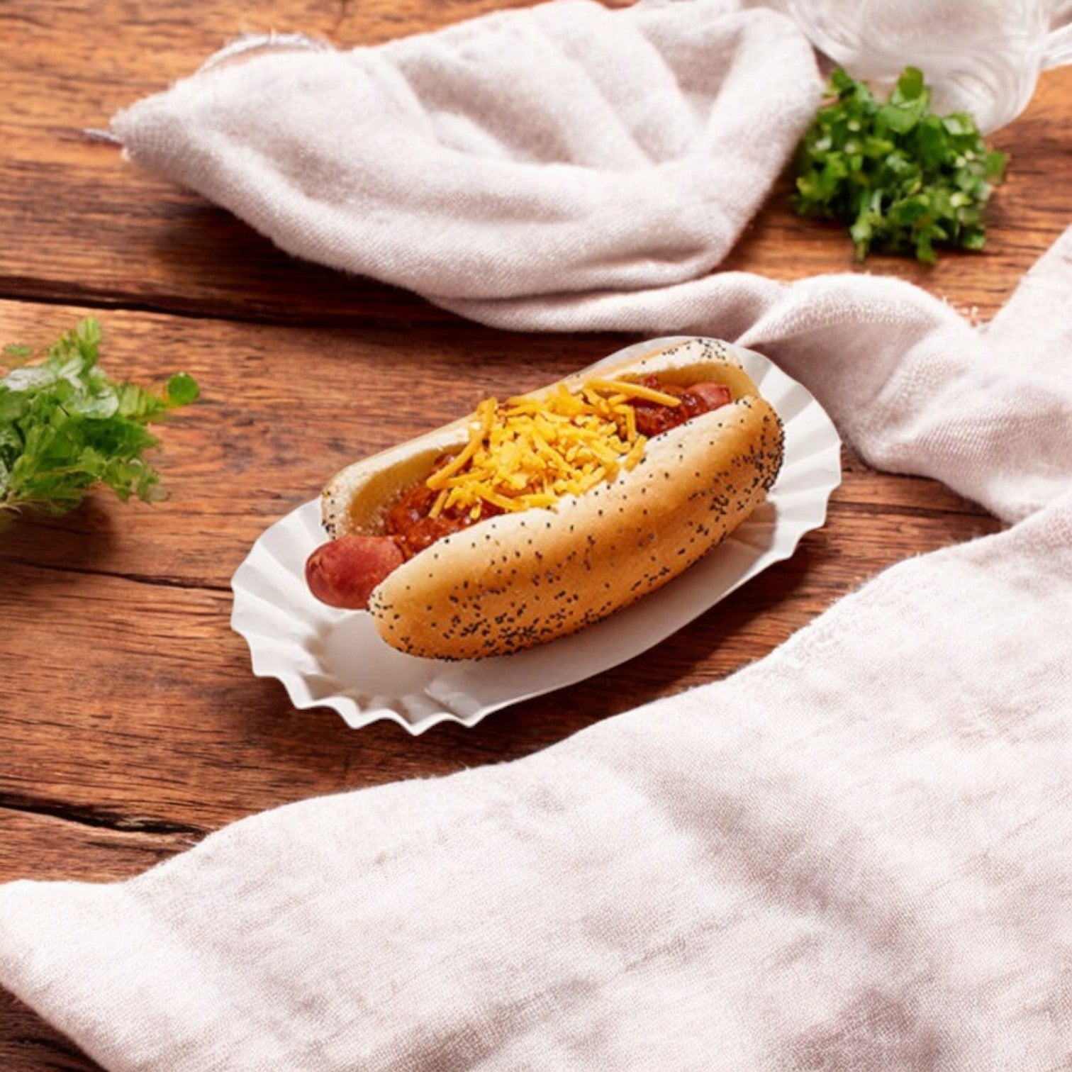 A Vienna Beef hot dog in a poppy seed bun, topped with shredded cheese and Bistro Soups by Vienna Beef – Chili Dog Chili, sits on a wooden table with a textured white cloth and parsley garnish.