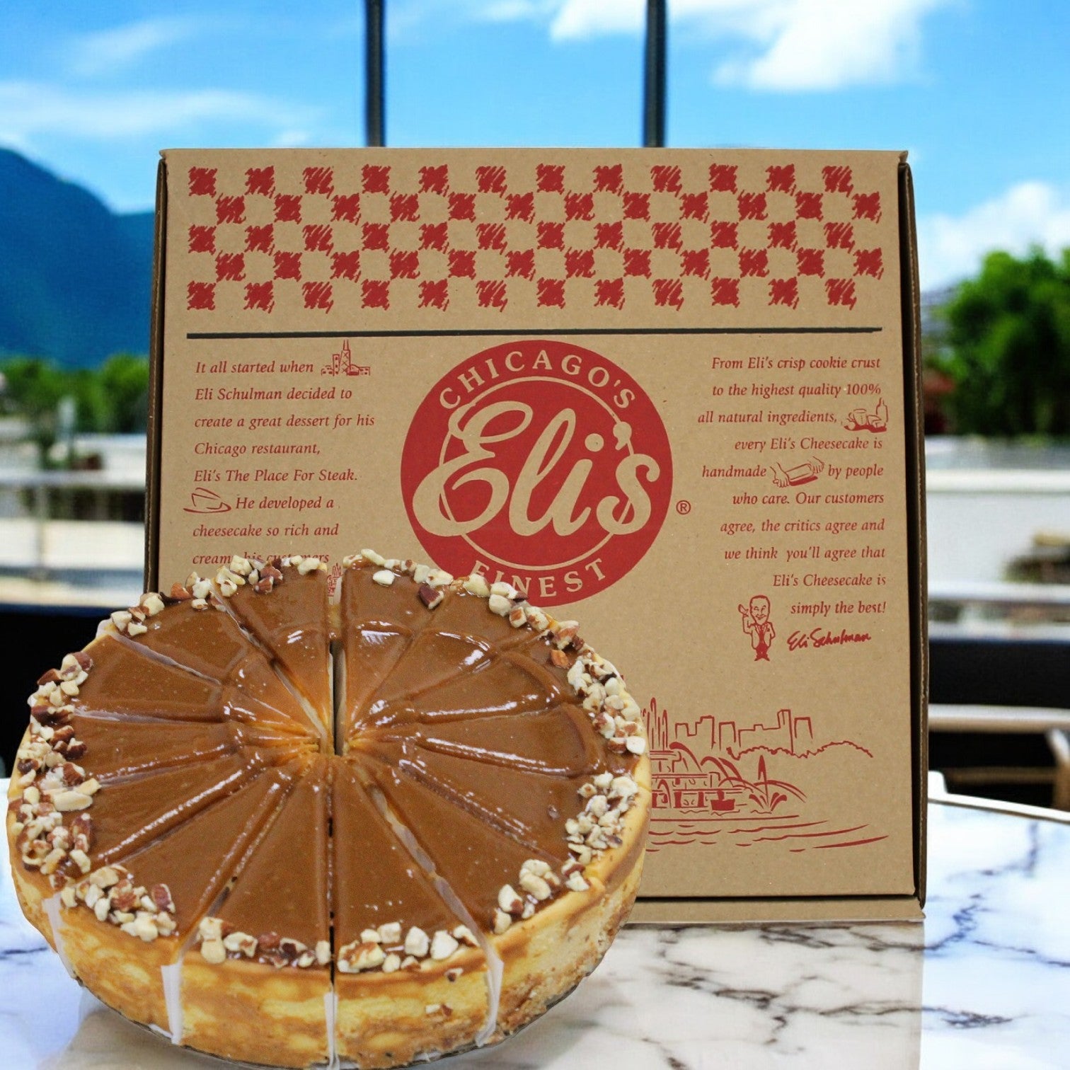 An indulgent Eli's Cheesecake Caramel Salted Cheesecake, topped with nuts, is elegantly placed on an outdoor table beside its box, surrounded by a scenic mountain view.