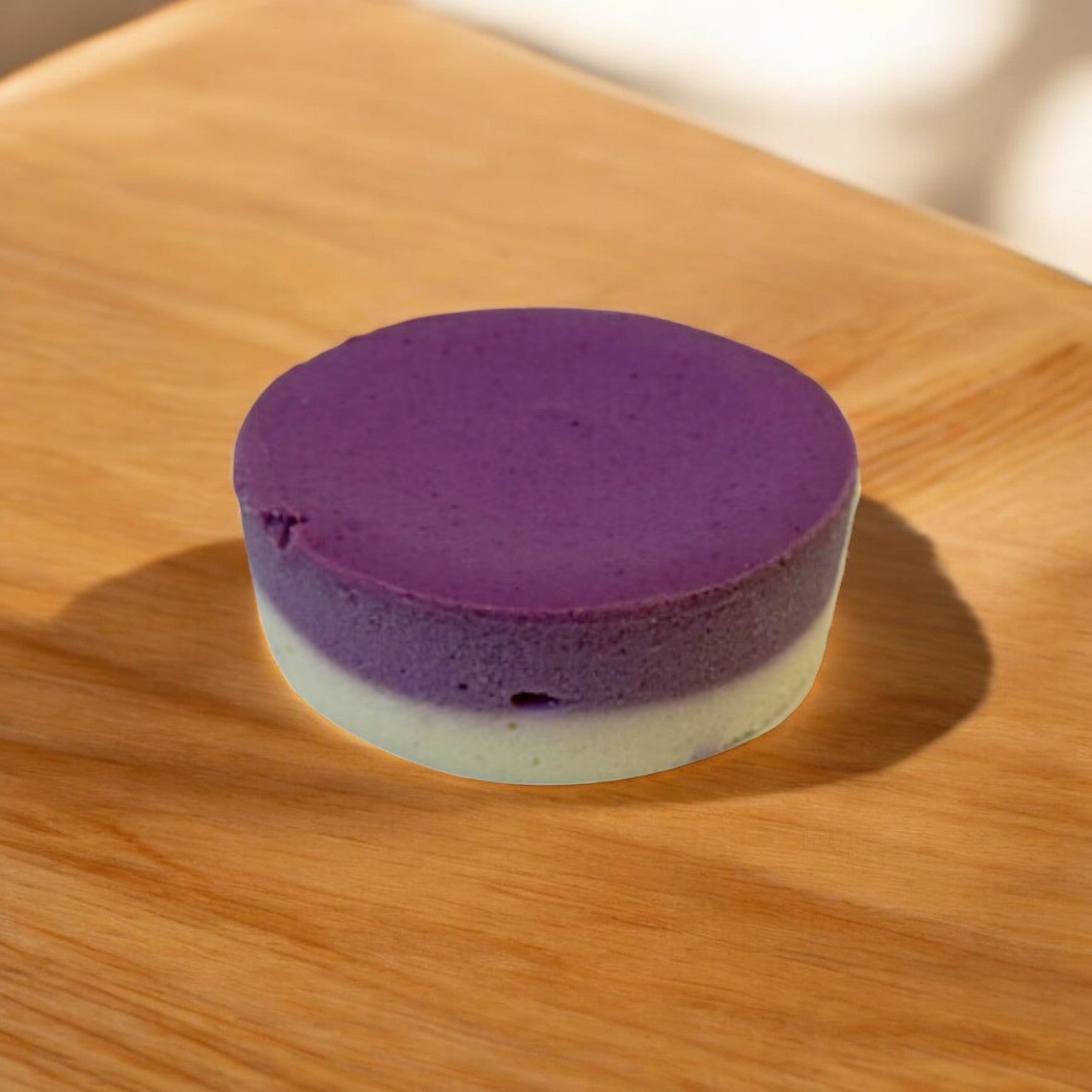 A Sweet Street Cheesecake Ube Individual, round and purple with cream highlights, rests on a wooden surface in partial shadow, crafted for a luxurious treat.