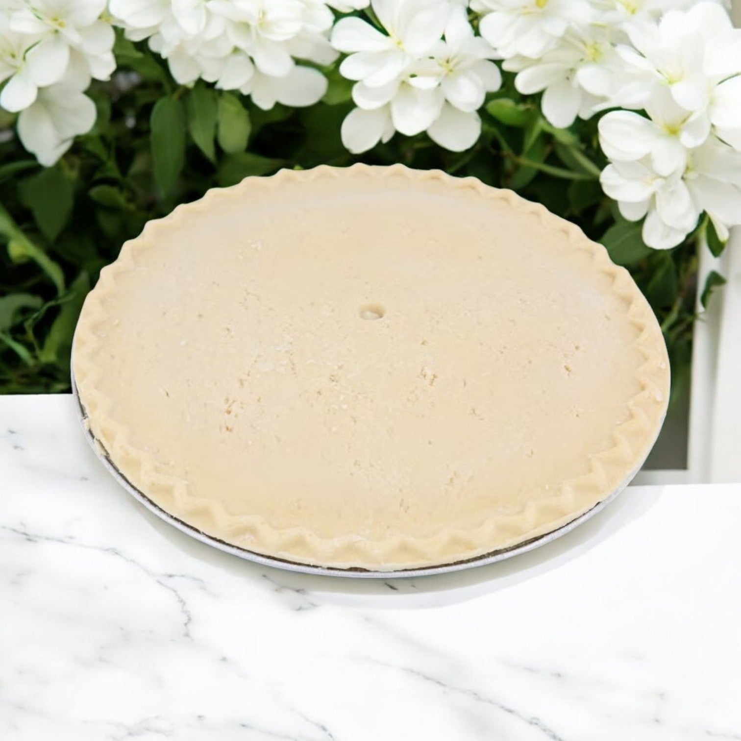 A Chef Pierre Peach Pie, weighing 46 oz, sits chilled in its tin on a marble surface adorned with delicate white flowers, ready to become a delicious masterpiece.