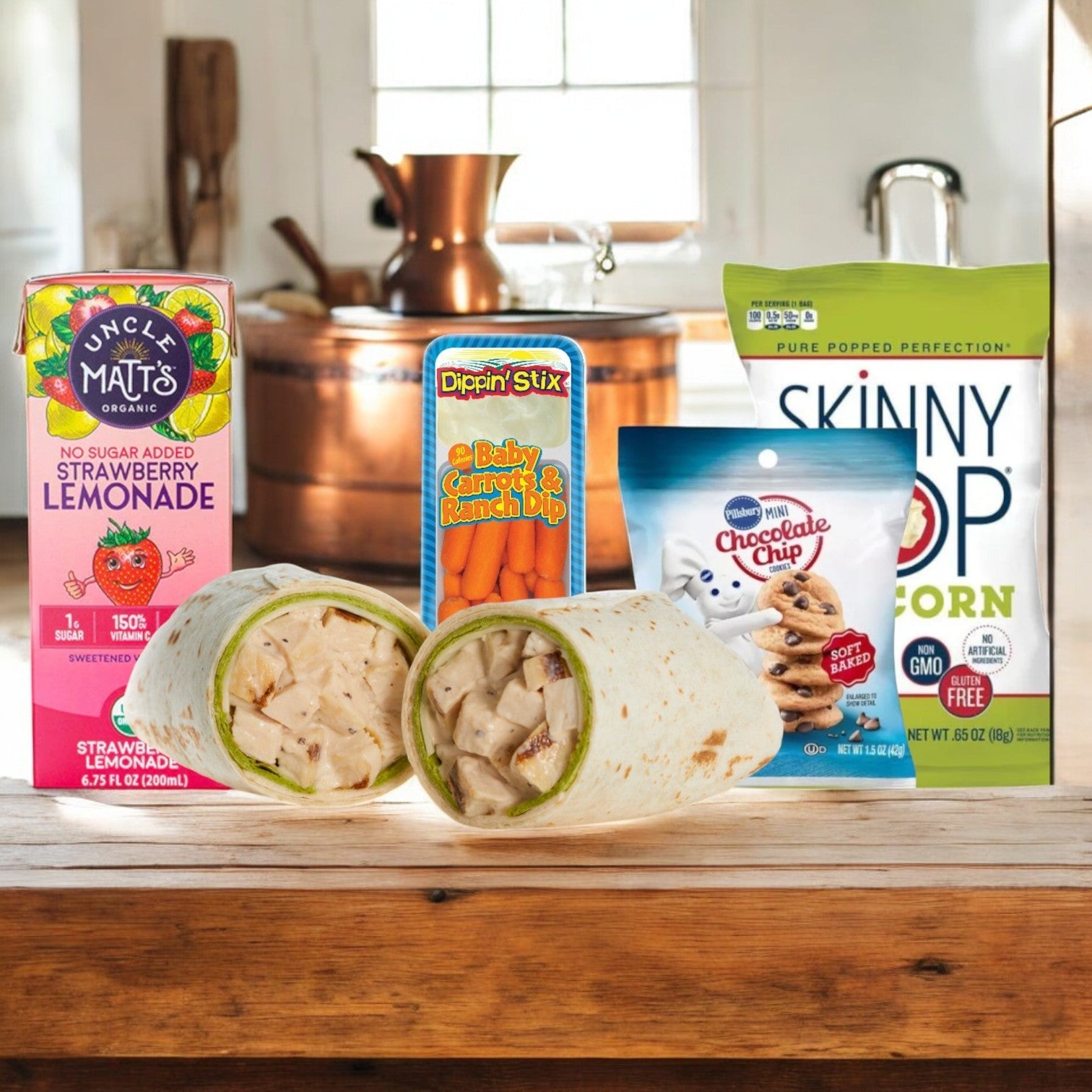 On a wooden table set, Easy Lunches' Chicken Caesar Wrap Lunch features a whole wheat wrap filled with creamy dressing, carrot sticks and dip, popcorn, refreshing lemonade, and chocolate chip cookies. Copper kitchenware subtly gleams in the background.
