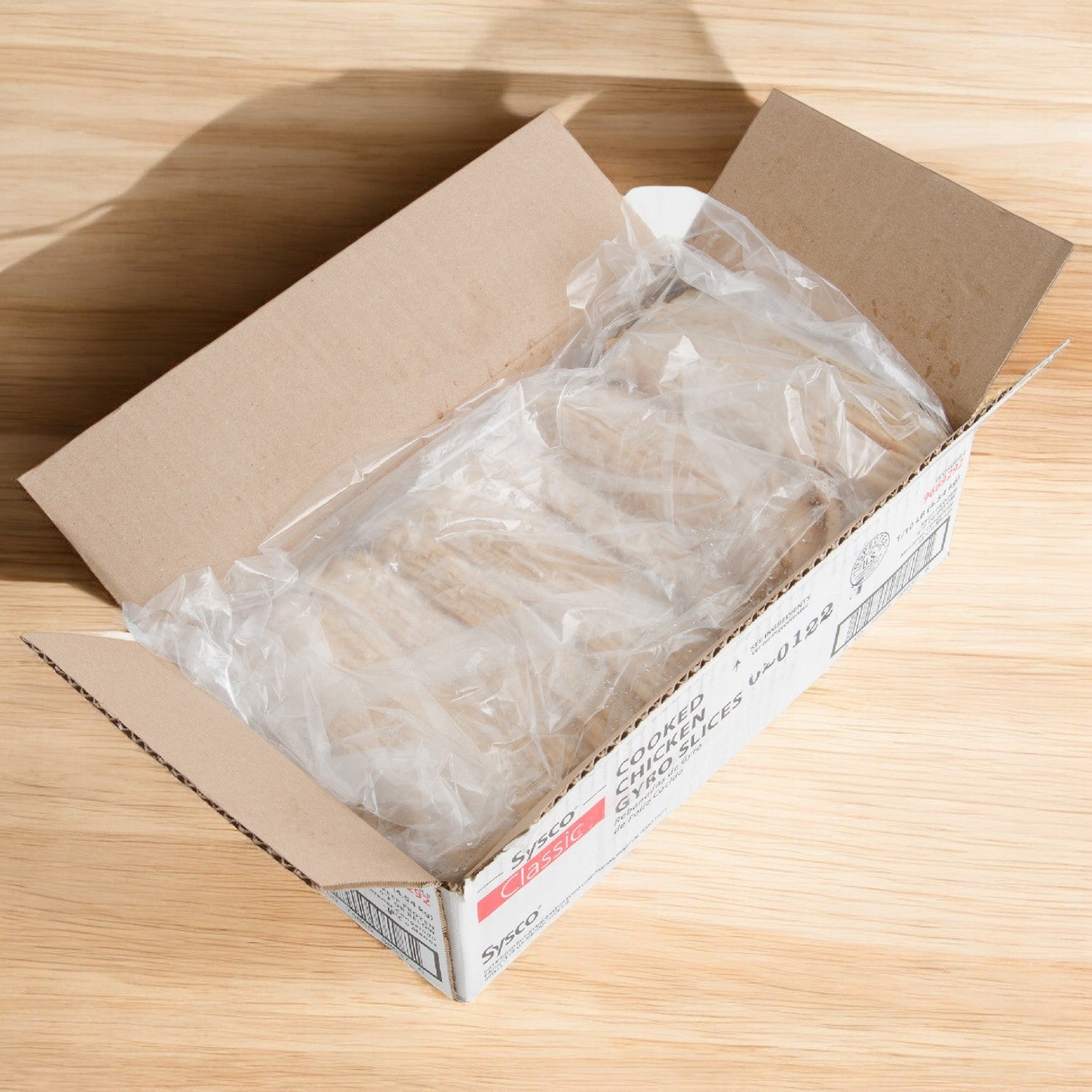 A cardboard box holds a plastic-wrapped Sysco Classic Chicken Gyro Meat Sliced Precooked, 10 lbs. - 1 Case on a wooden surface, ready to bring the authentic Mediterranean taste to your table.