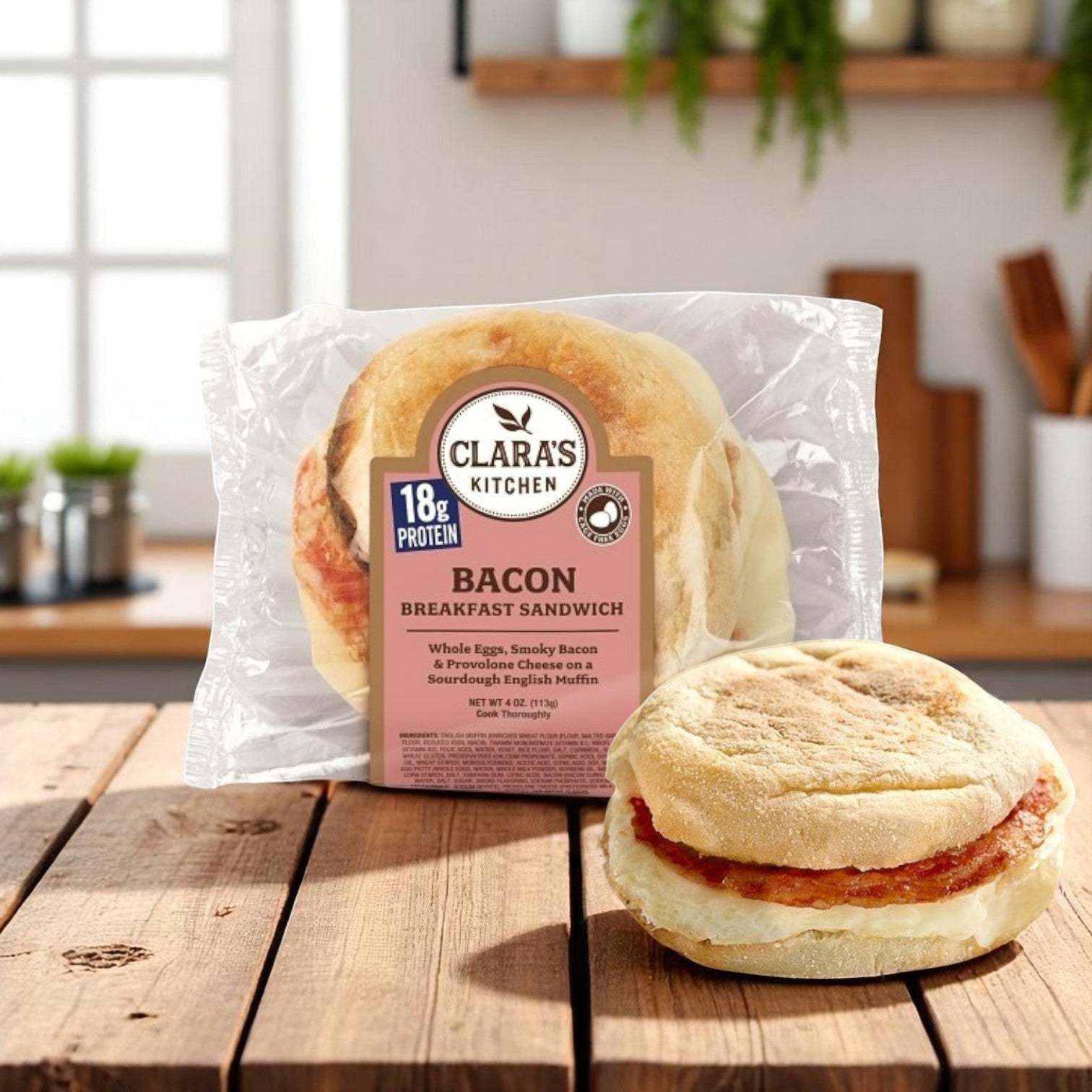 Clara's Kitchen Individually Packaged Bacon Breakfast Sandwich, 4 oz., sits on a wooden table with an unwrapped one beside it. Kitchen items and a plant create a gently blurred backdrop.