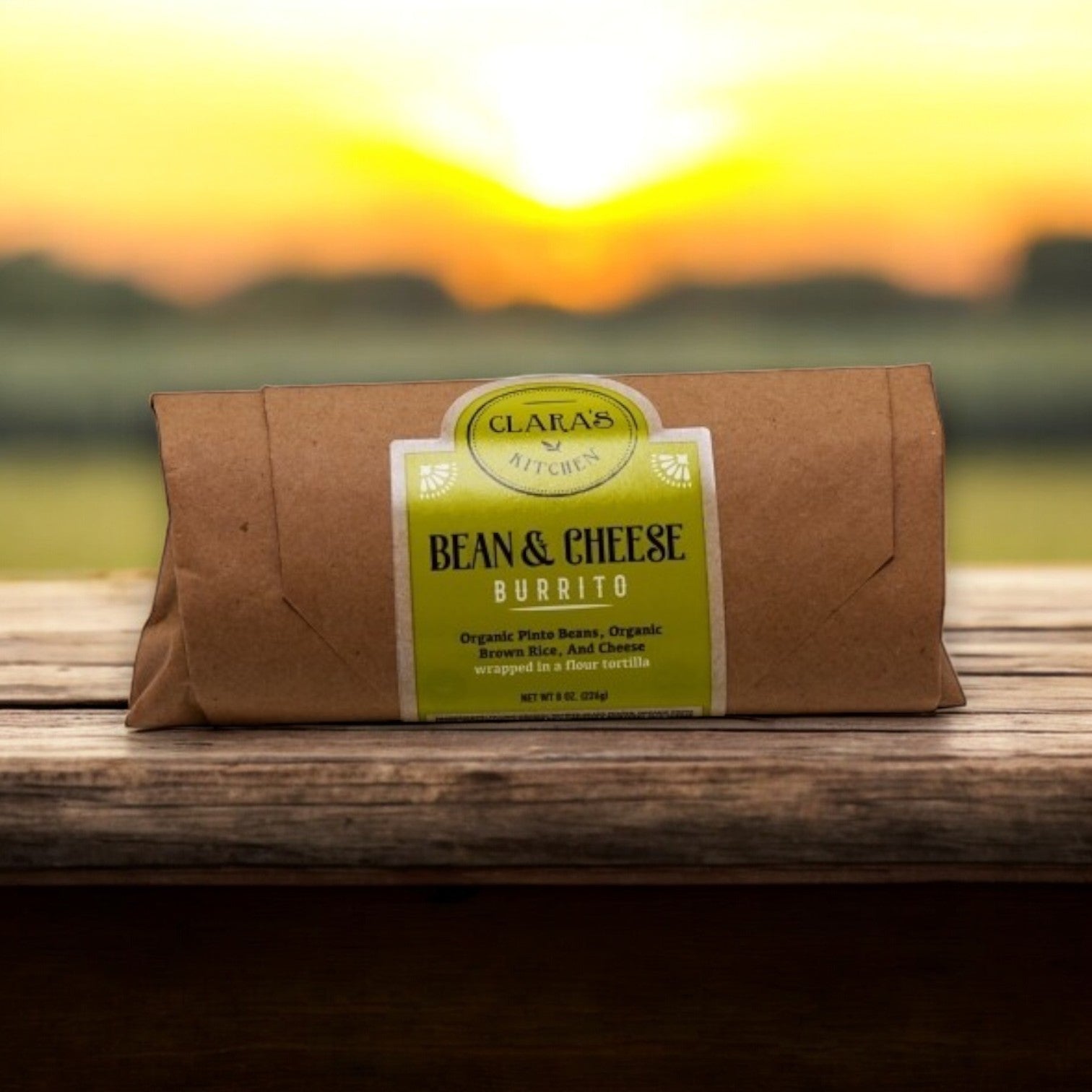 An 8 oz. Clara's Kitchen Individually Packaged Bean & Cheese Burrito lies on a wooden surface, with a stunning blurred sunset in the background.