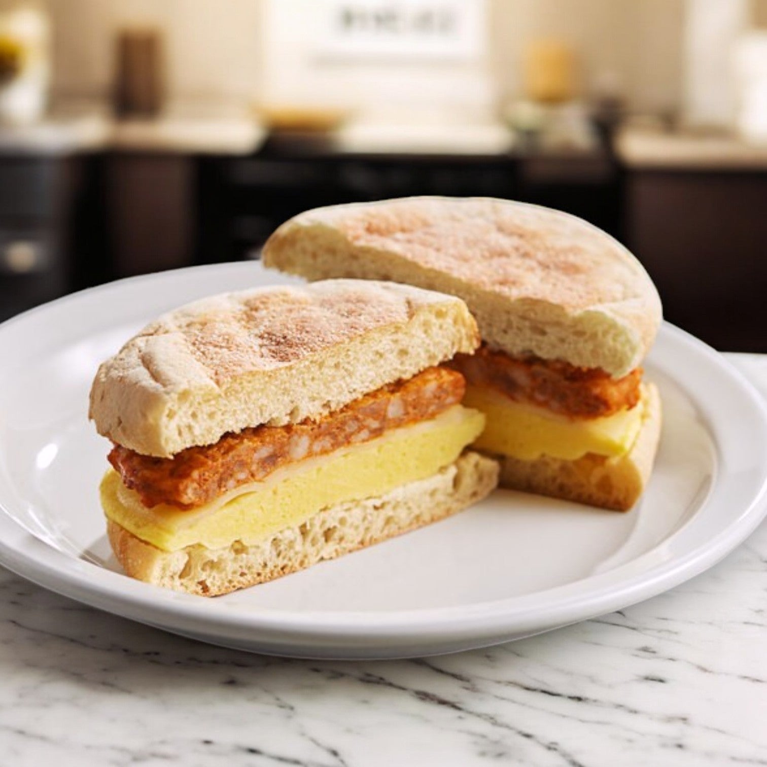 Start your day with Clara's Kitchen Chorizo Breakfast Sandwich. Served on an English muffin with egg and cheese, it includes a crispy chorizo patty. Individually packaged for convenience, this 5oz sandwich is halved on a white plate in a kitchen setting.