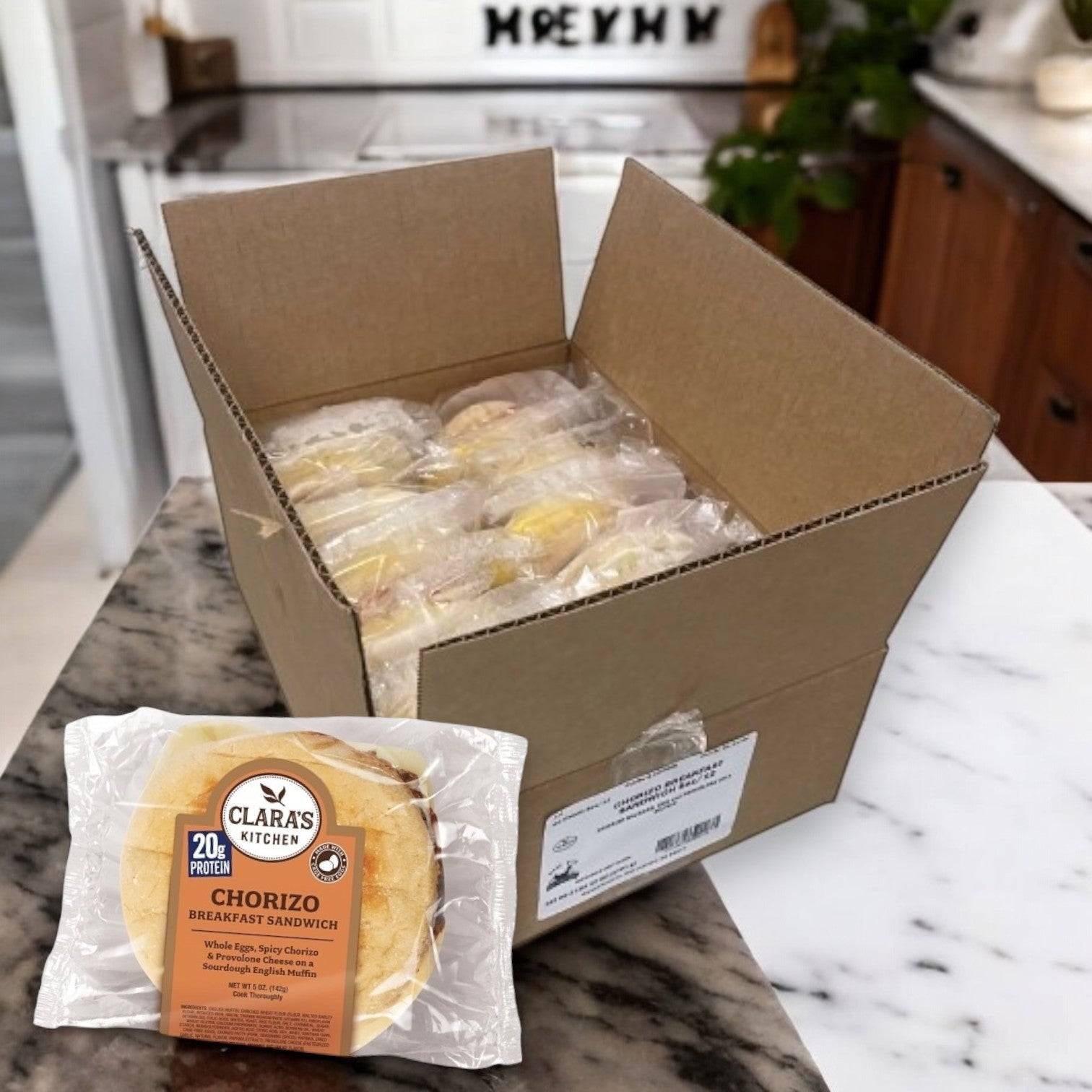 An open box of Clara's Kitchen Chorizo Breakfast Sandwiches, each 5oz., sits on a marble kitchen counter. These individually packaged, convenient breakfast choices include a round bun and protein label.