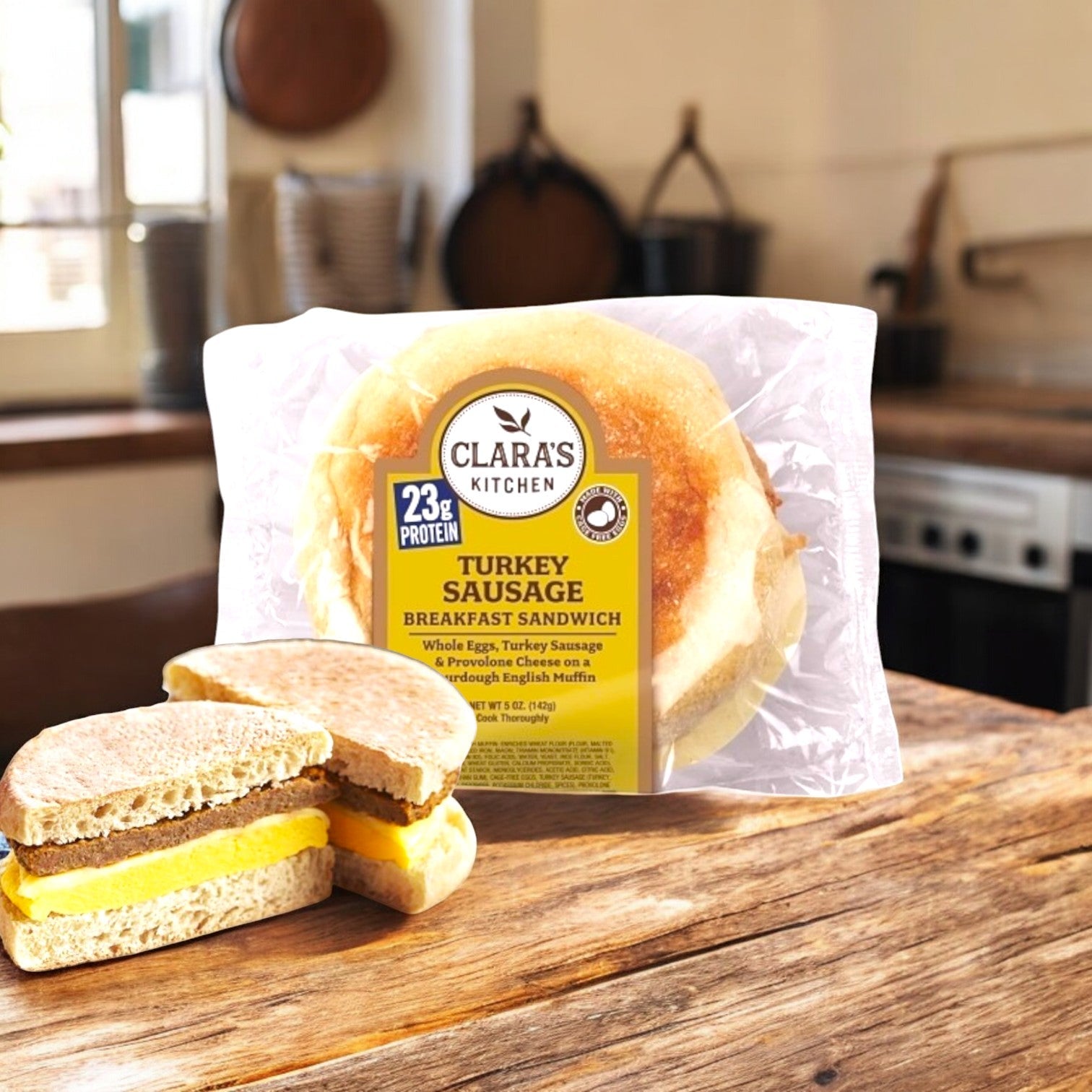 Clara's Kitchen's 5 oz. Turkey Sausage Breakfast Sandwich, individually packaged, sits on a wooden table. In front, another enticing sandwich, served on an English muffin and sliced in half, complements a cozy kitchen setting.