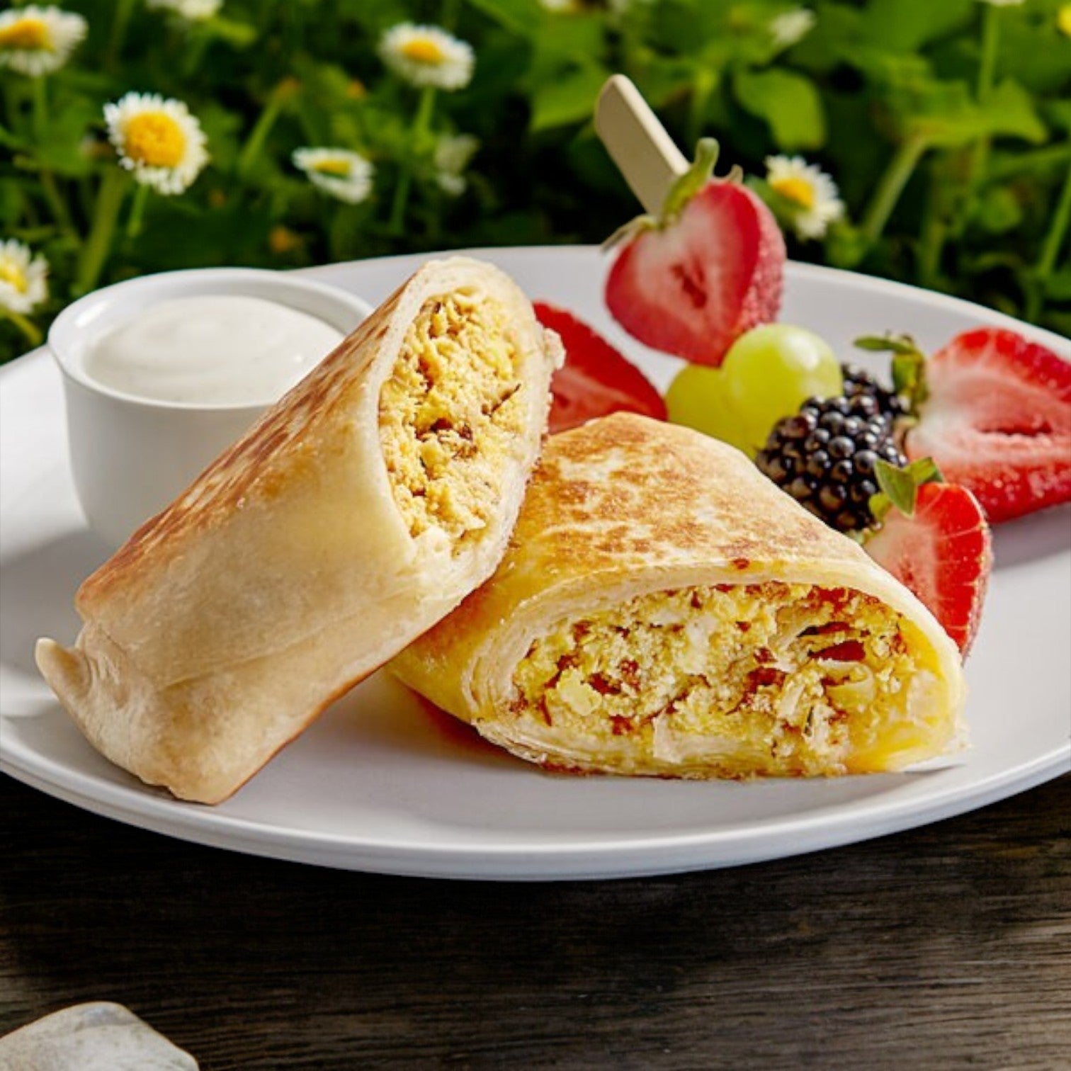Clara's Kitchen Individually Packaged Sausage Breakfast Burrito is halved on a white plate with mixed berries and sauce, set against a lush green backdrop—ideal as a grab-and-go option for busy mornings.