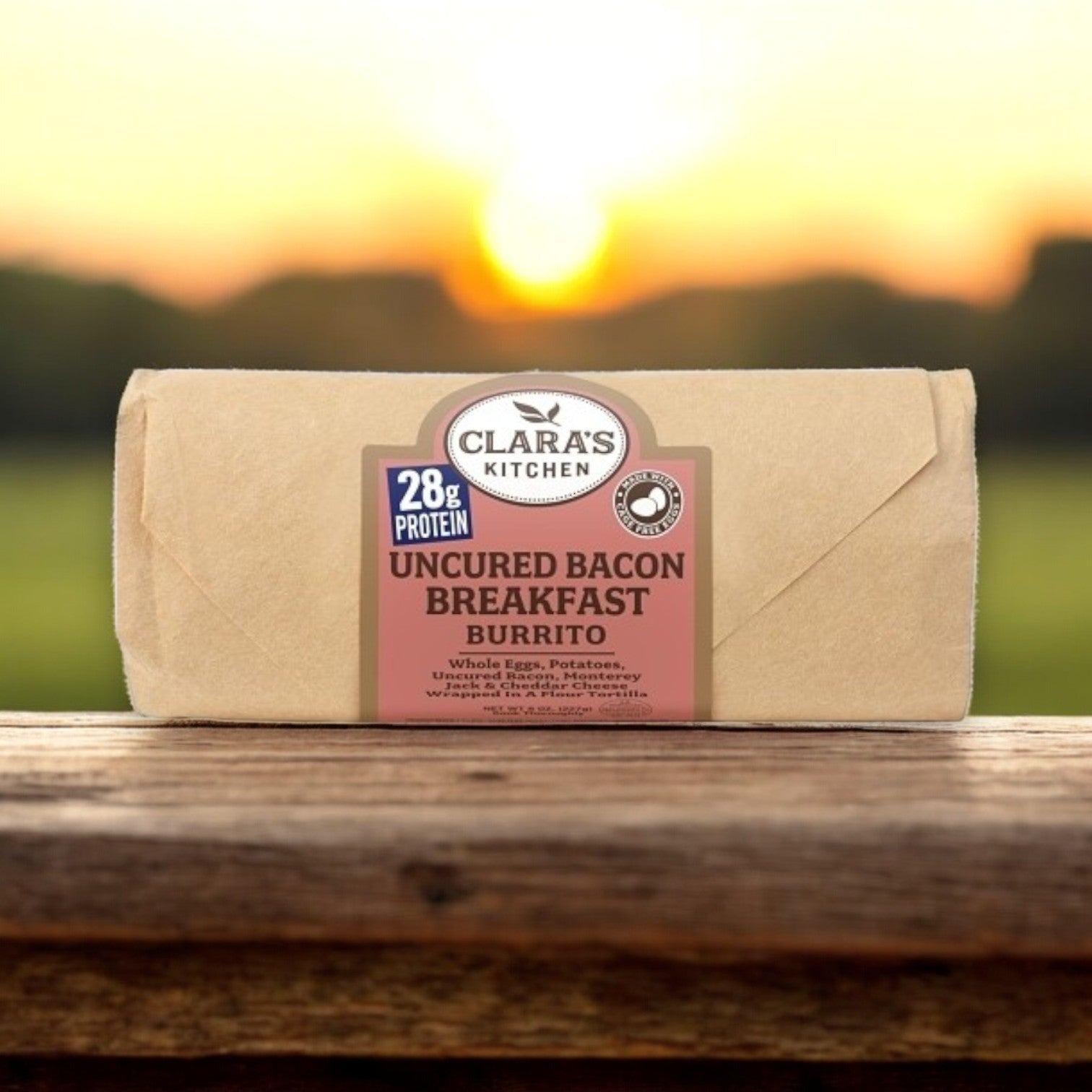 Clara's Kitchen Individually Packaged Uncured Bacon Breakfast Burrito, weighing 8 oz., sits on a wooden table with a blurred outdoor sunrise backdrop, offering breakfast lovers a convenient choice.