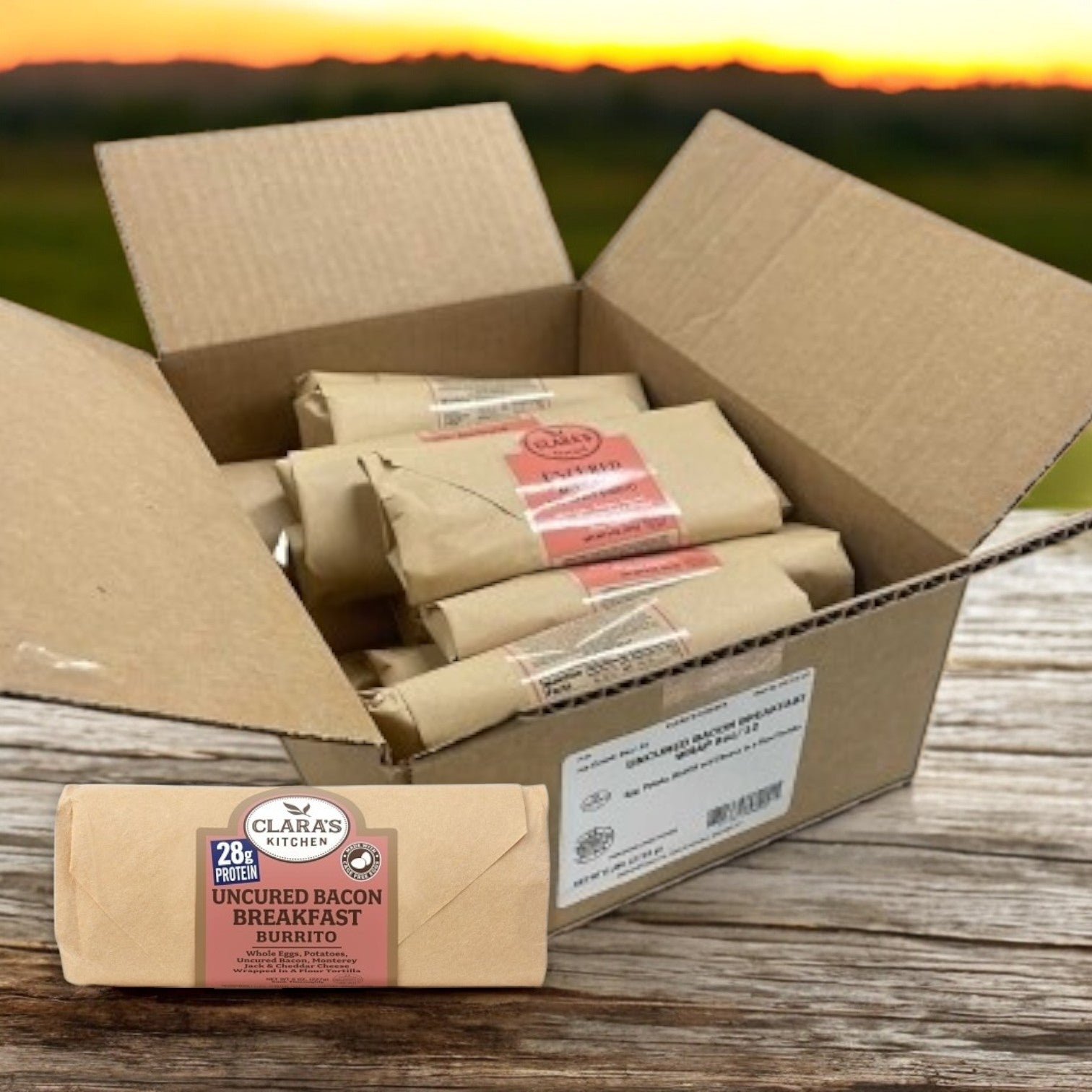 An open box of Clara's Kitchen Individually Packaged Uncured Bacon Breakfast Burritos sits on a wooden surface. One burrito lies in front, against a stunning sunset, offering a convenient meal for any time of the day.