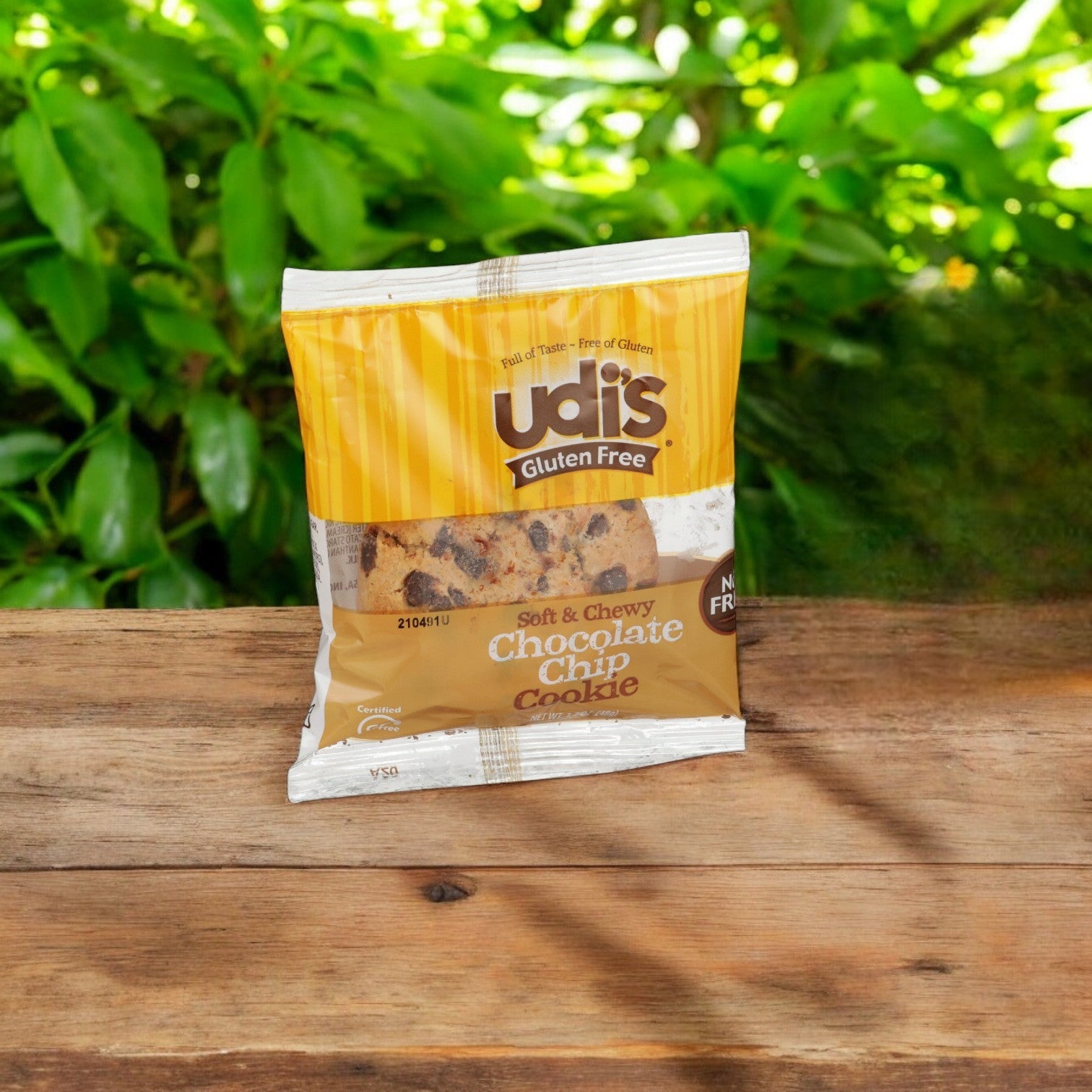A package of Udi's Gluten Free Chocolate Chip Cookies, available in a case of 36, rests on a wooden surface with green foliage in the background.
