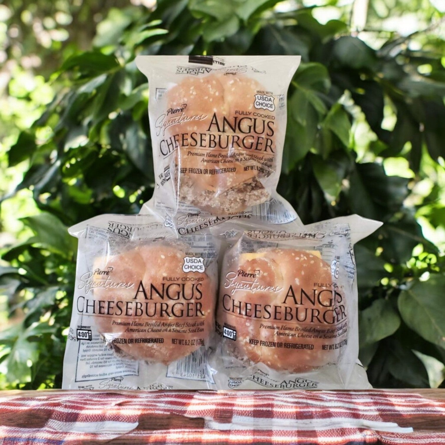 Three Tyson Pierre Signature Angus Cheeseburgers from the 8 Count, 3 Pack are stacked on a red checked tablecloth outdoors, with green foliage in the background.