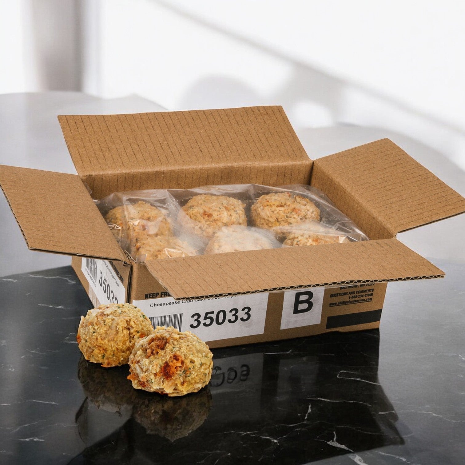 A Phillips Foods box holds plastic-wrapped cheese biscuits beside a delectable 3.25 oz. Crab Cake Chesapeake on a black marble surface, with two biscuits outside.