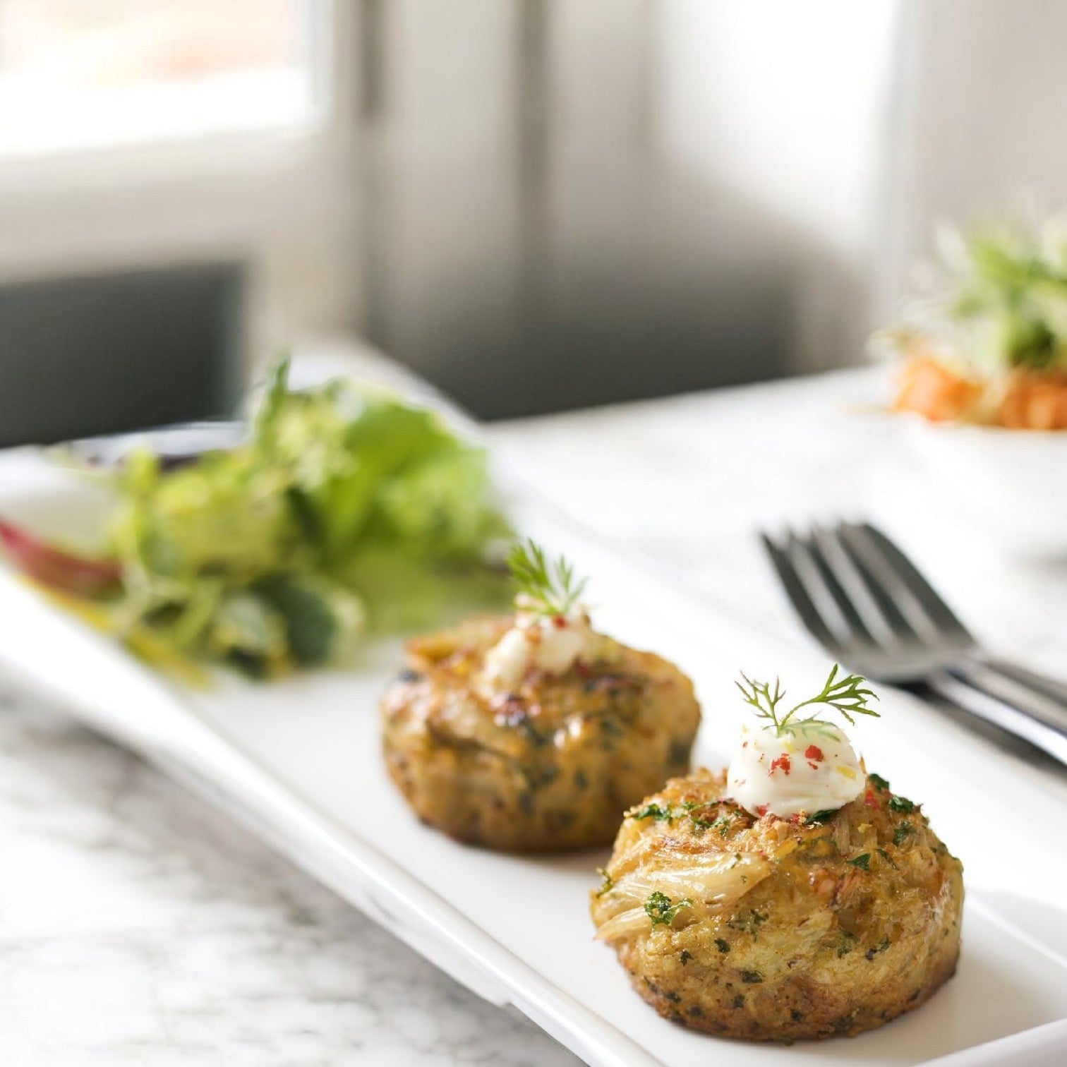 Two 3.25 oz. Phillips Foods Chesapeake Bay Crab Cakes are topped with sauce and herbs on a white rectangular plate, accompanied by mixed greens.