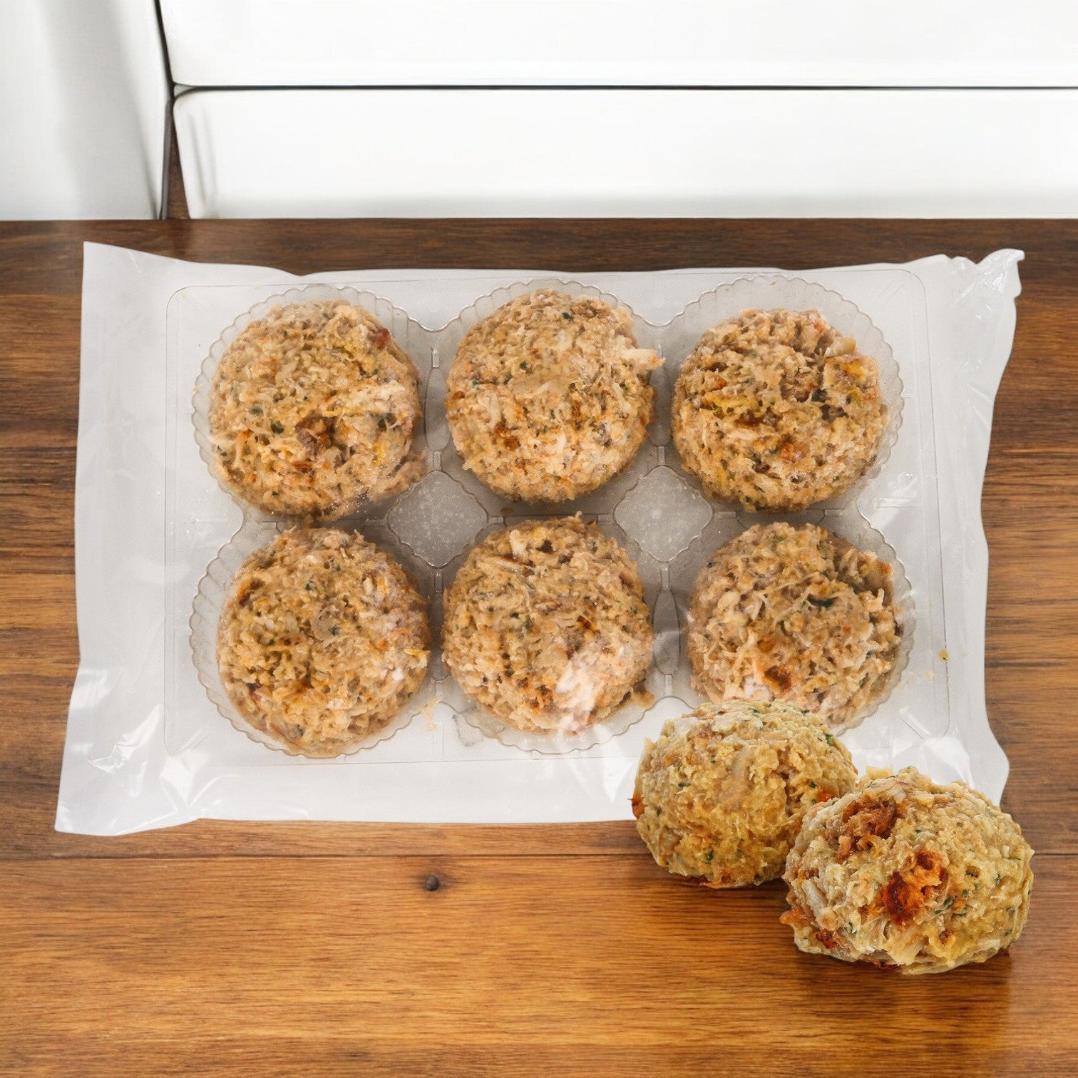 A sealed tray of Phillips Foods Crab Cake Chesapeake holds six stuffing balls on a wooden surface, reminiscent of Chesapeake Bay, with two more in front.