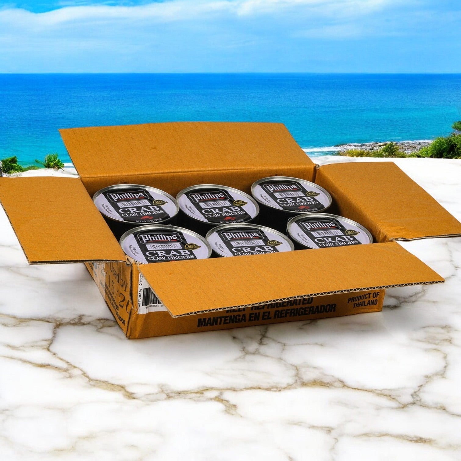 An open cardboard box reveals several cans of Phillip Foods Crab Claw Finger Pasteurized Asia, 12 oz., by Phillips Foods on a marble surface against an ocean view, embodying the essence of sustainable seafood delights.