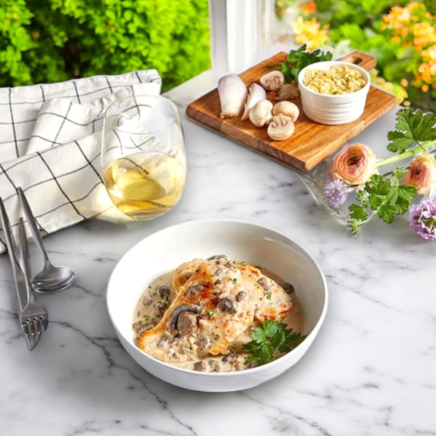 On a marble table sits a bowl of Three Little Pigs – Sous-Vide Creamy Chicken with Mushrooms. A glass of white wine is nearby, along with a napkin with cutlery and a cutting board filled with premium ingredients. Flowers and green foliage enhance the charming backdrop.