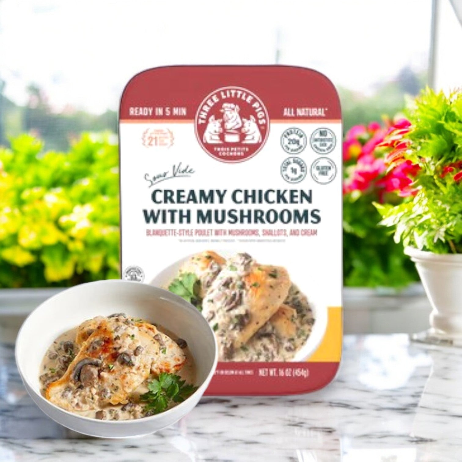 A 16 oz package of Three Little Pigs – Sous-Vide Creamy Chicken with Mushrooms by Trois Petits Cochons sits on a table behind a bowl of the finished dish in creamy sauce, with a blurred outdoor setting highlighting its premium ingredients.