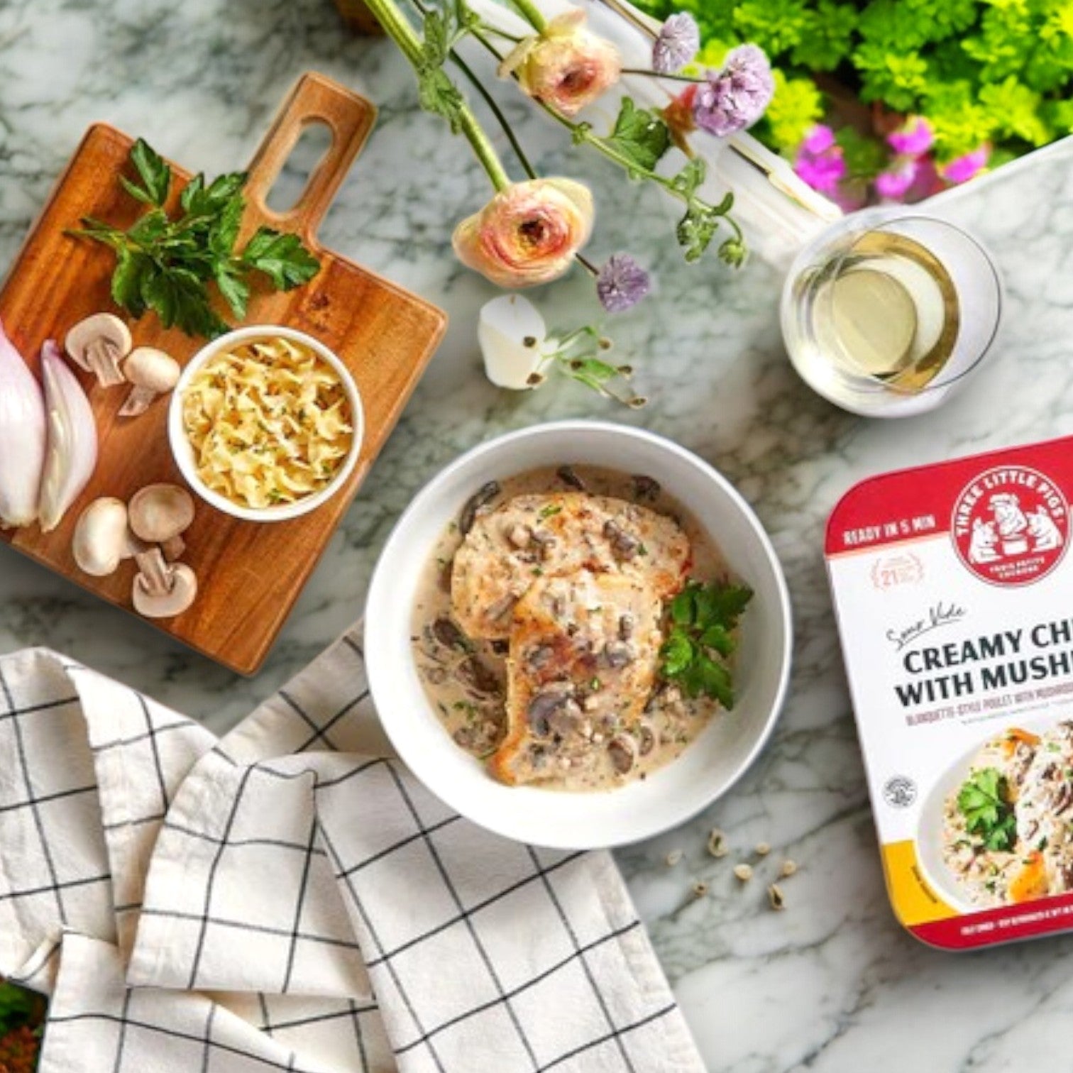 On a marble countertop sits a bowl of creamy chicken and mushrooms, crafted from premium ingredients. Nearby, a cutting board with fresh herbs and pasta completes the scene, along with Three Little Pigs – Sous-Vide Creamy Chicken with Mushrooms (16 oz), delicate flowers, and a glass of white wine.
