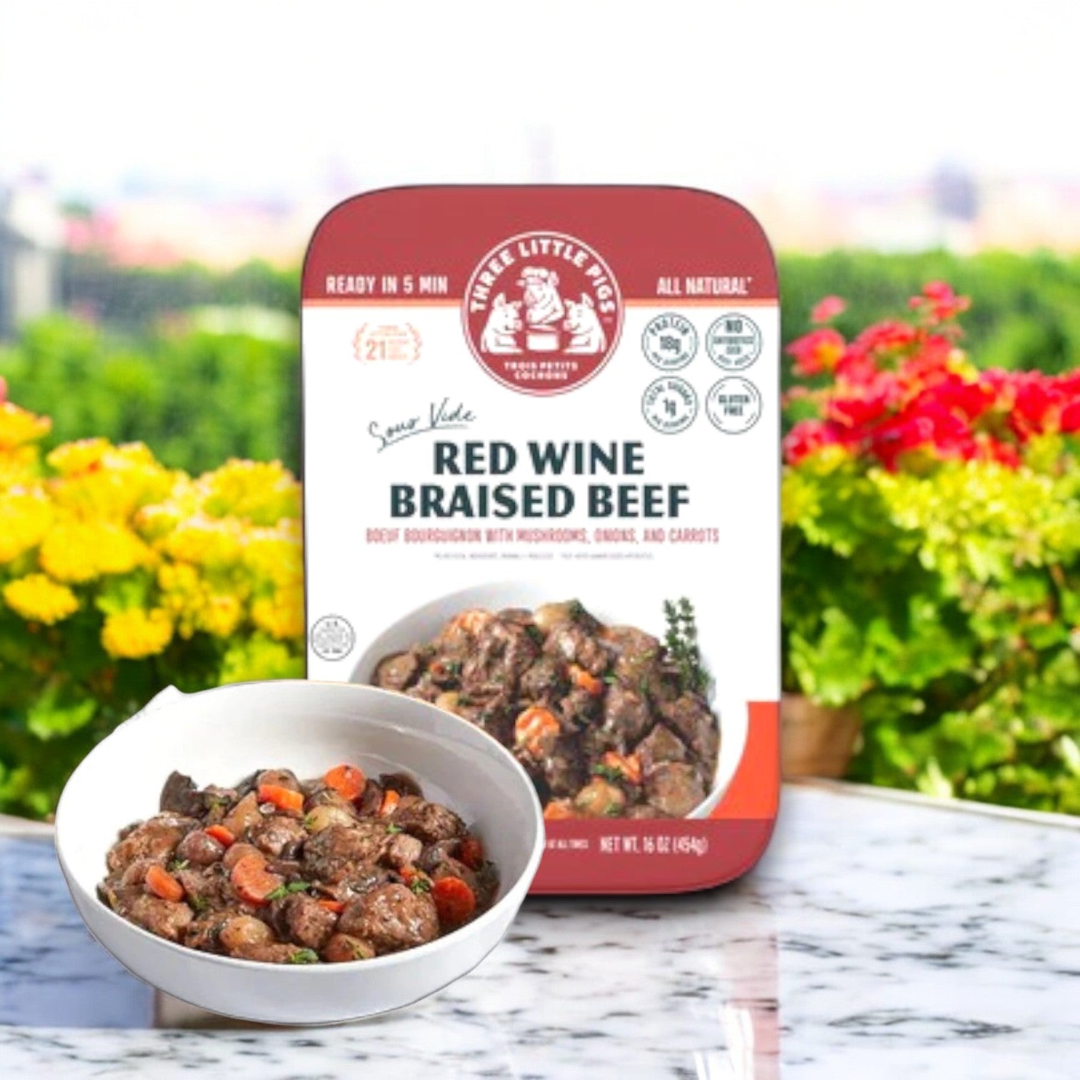 A 16 oz. container of Three Little Pigs – Sous-Vide Red Wine Braised Beef sits on the table, offering a restaurant-quality experience against a charming garden view. Crafted by Trois Petits Cochons, it promises an elevated dining indulgence with premium ingredients and vegetables.