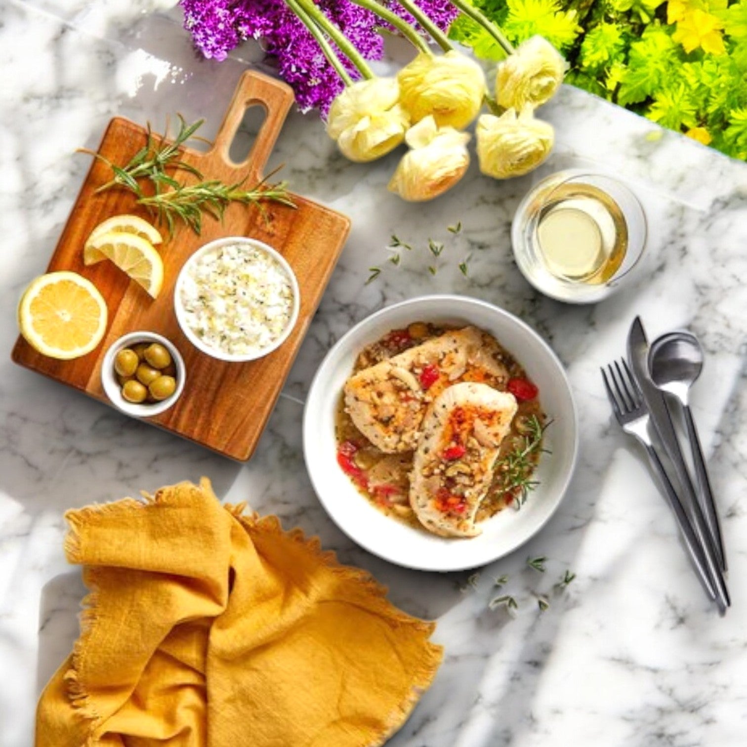 A bowl of Three Little Pigs – Sous-Vide Lemon & Herb Chicken is on a marble table, accompanied by lemon, olives, extra sauce on a cutting board, yellow cloth, cutlery, white wine glass, and vibrant flowers. Premium ingredients from Three Little Pigs Trois Petits Cochons enhance this elegant meal.