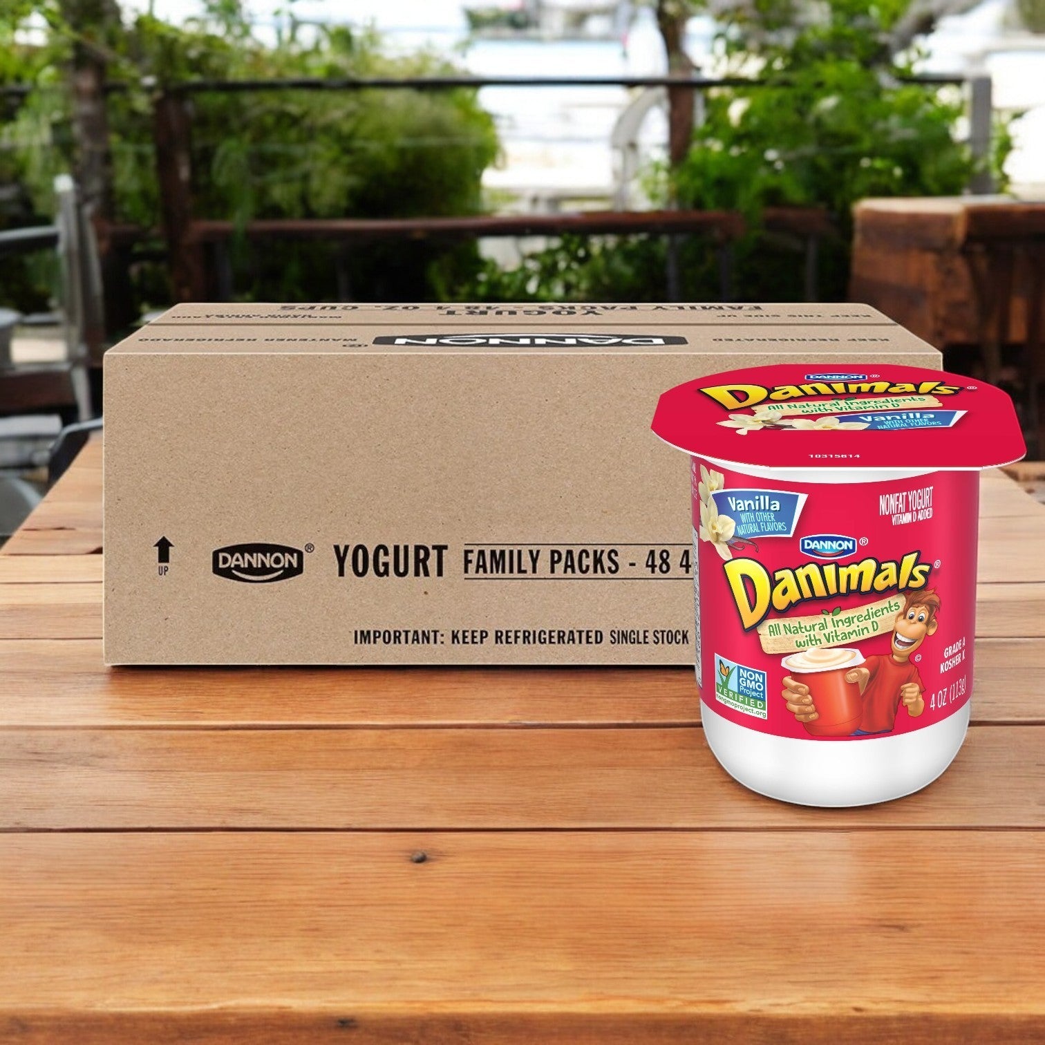 A large cardboard box labeled "Danimals Vanilla Nonfat Yogurt, 4 oz. Dannon - 48 per Case" sits on a wooden table next to a cup of kid-friendly vanilla yogurt.