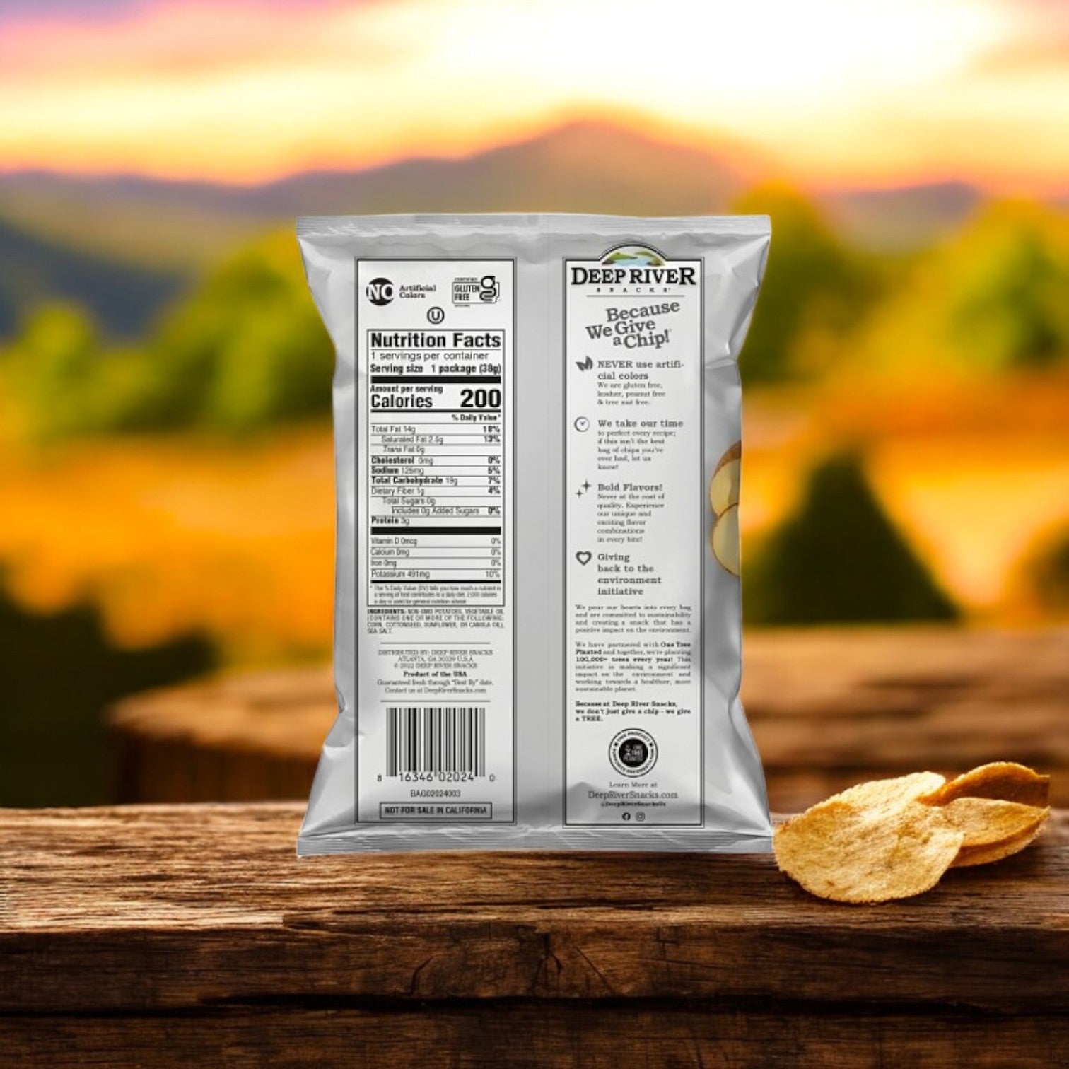 A bag of Deep River Snacks Original Sea Salt Kettle Potato Chips, non-GMO and gluten-free, is on a wooden surface with some chips scattered around. Behind it is a blurred scenic sunset over hills.