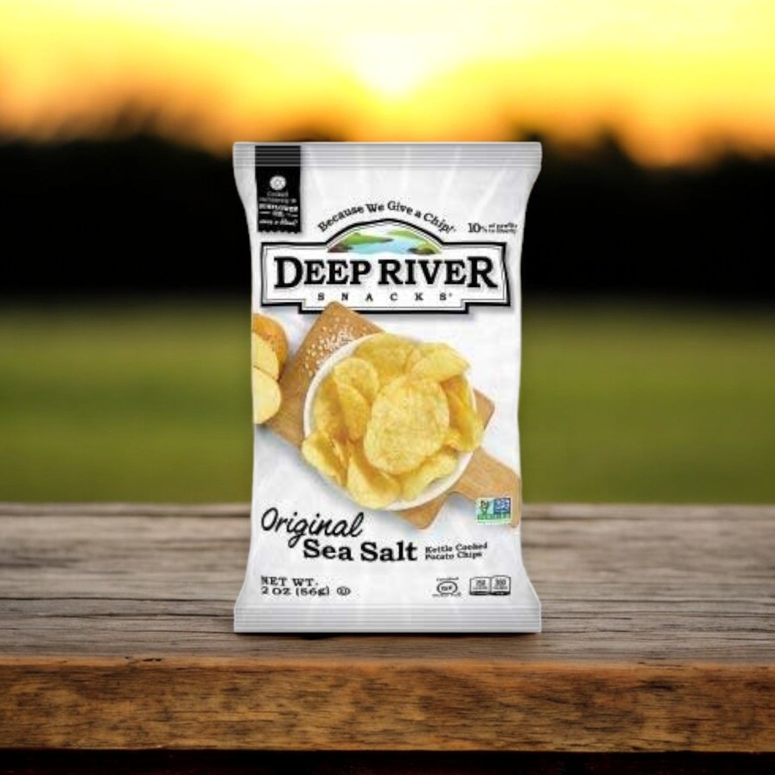 A bag of Deep River Snacks - Original Sea Salt Kettle Potato Chips (gluten-free, kosher, 2 oz) is on a wooden surface with a blurred sunset in the background.