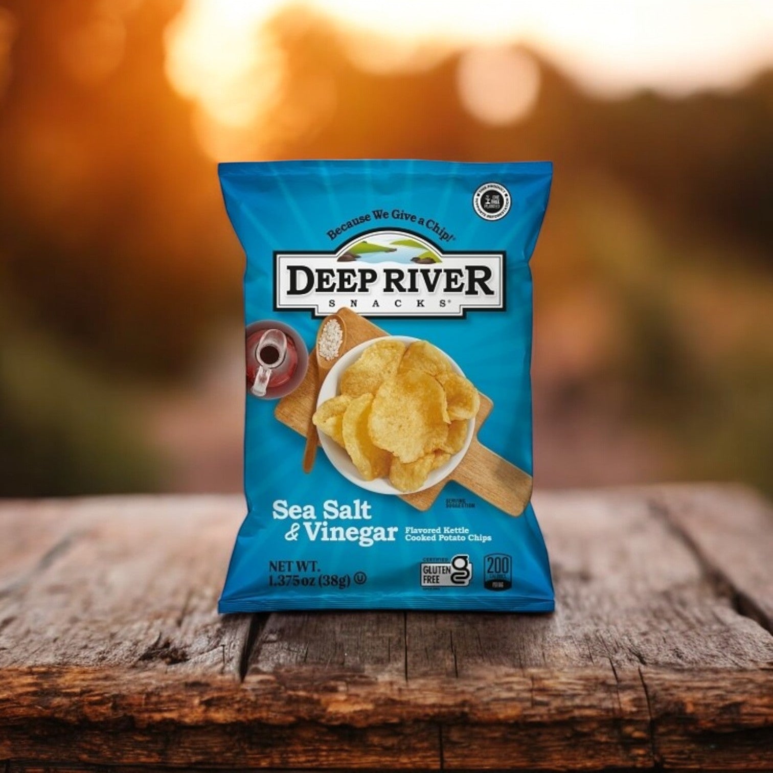 A 1.375-ounce bag of Deep River Snacks Salt & Vinegar Kettle Potato Chips, Non-GMO and Gluten-Free, is displayed on a wooden surface with a blurred outdoor backdrop.