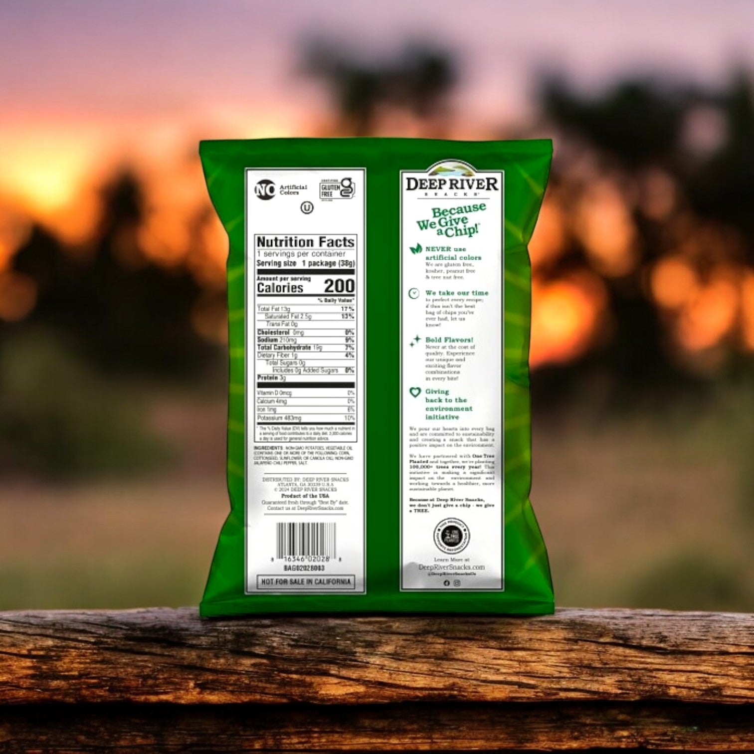 The back of a green bag of Deep River Snacks Zesty Jalapeno Kettle Potato Chips includes nutritional details. These gluten-free chips offer a flavorful punch, set against a blurred sunset background.