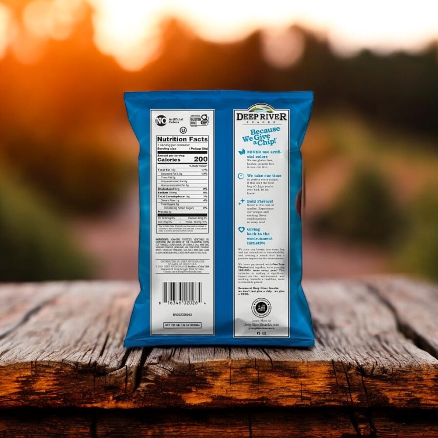 A bag of Deep River Snacks Salt & Vinegar Kettle Potato Chips sits on a wooden table, highlighting its Non-GMO and Gluten-Free labels. The out-of-focus outdoor setting in the background adds to the natural charm.