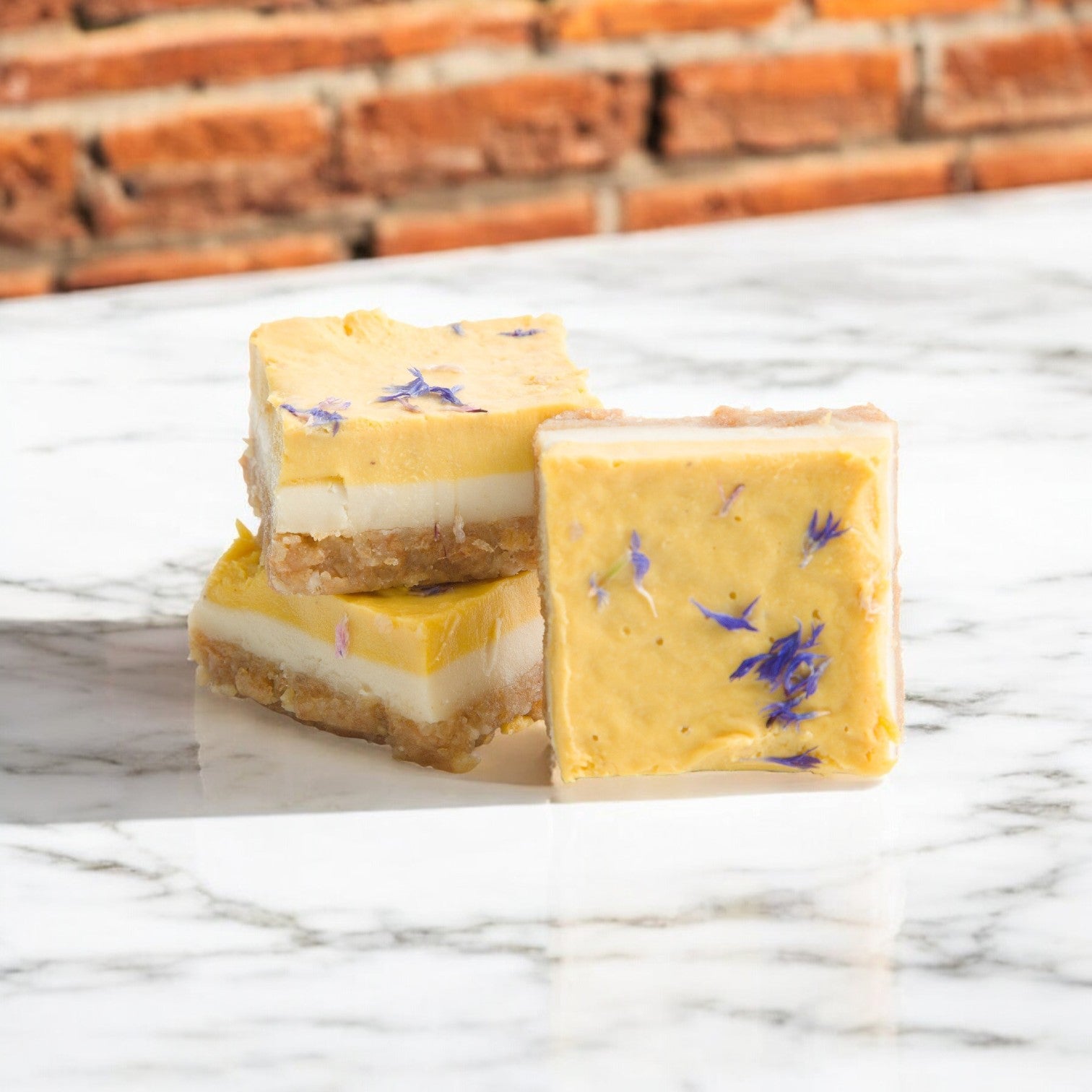 Three Nats Vegan Cakes square dessert bars with layered lime mango filling and blue flower petals rest on a marble surface against a brick wall. This vibrant, plant-based treat is both inviting and delicious.