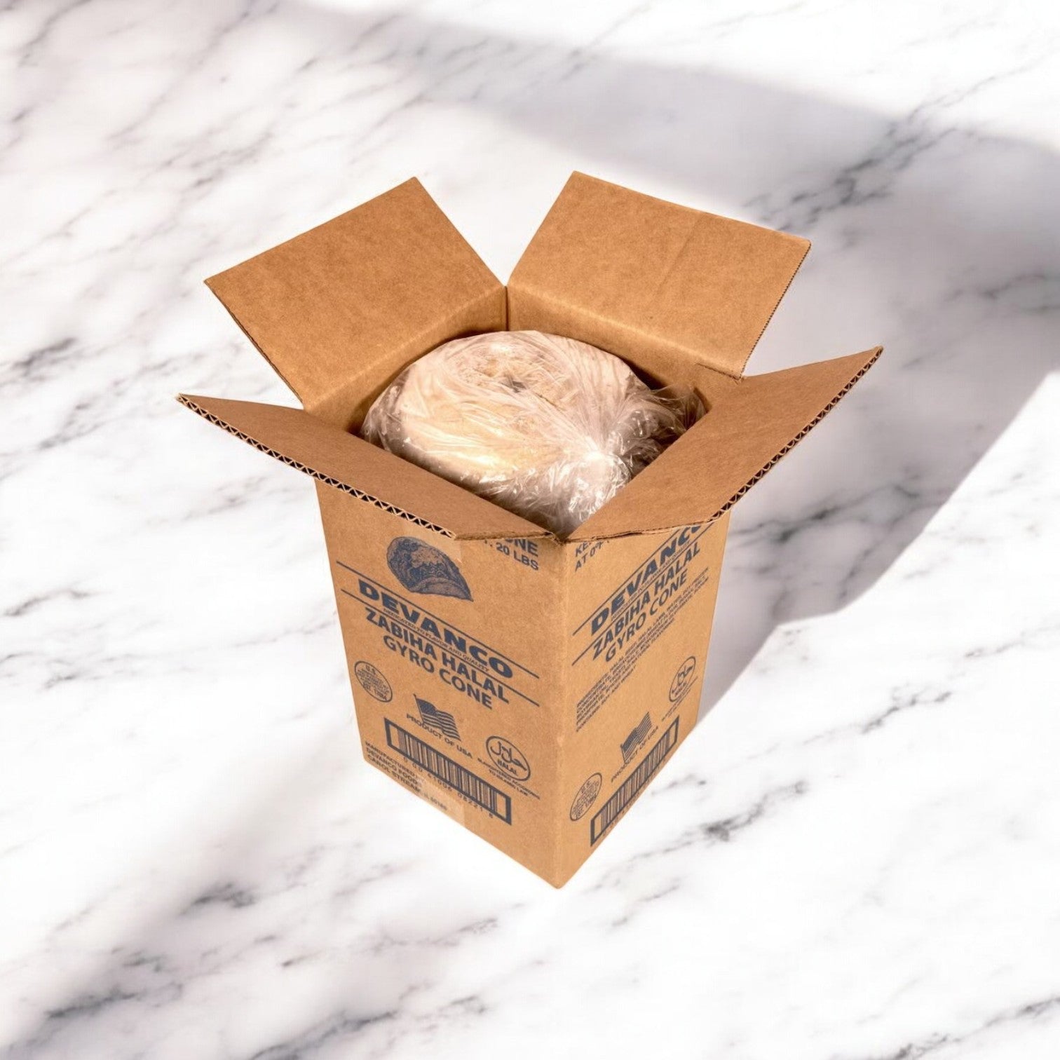 An open cardboard box on a marble surface reveals a plastic-wrapped treasure, hinting at authentic Mediterranean flavor. Could it be Devanco Foods' Halal Off the Cone Gyro Slices waiting to delight?.