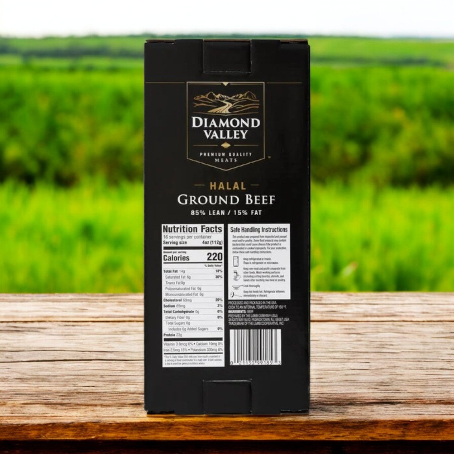 A black package of New Zealand Spring Lamb's Diamond Valley Halal Ground Beef, 85% lean, 15% fat, with nutrition facts and handling instructions, rests on a wooden surface against a blurred green field background. This premium product ensures high standards for your meals.