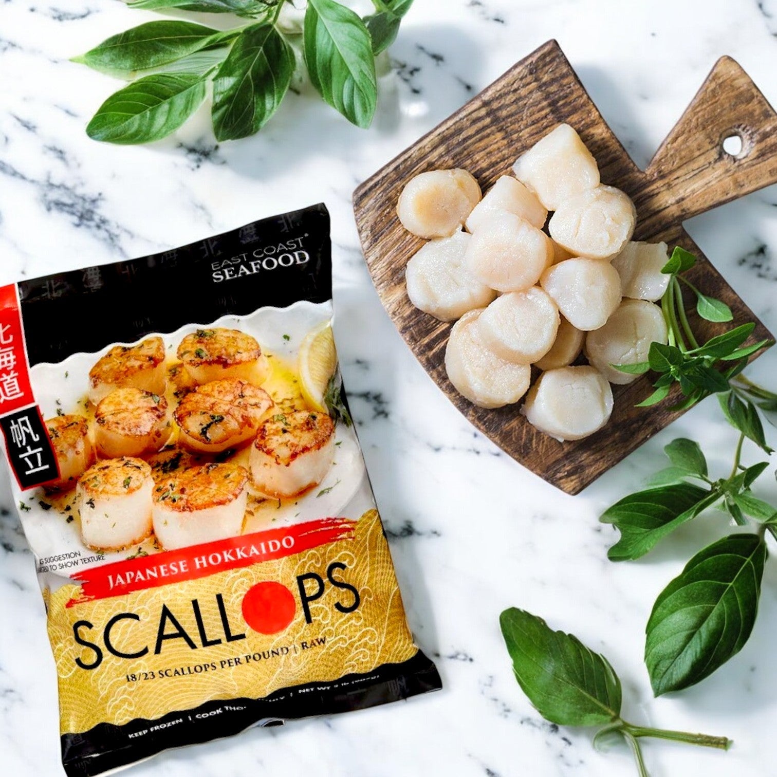 A 2-lb pack of Kirkland Signature East Coast Seafood Japanese Hokkaido Scallops (18-23 ct, raw) is shown beside a wooden board with raw seafood and fresh basil on a marble surface.