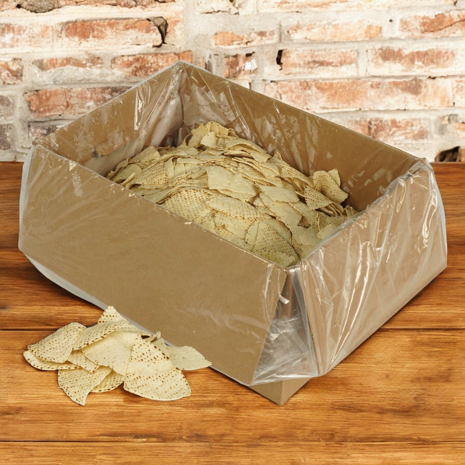 A large cardboard box lined with plastic holds bulk El Milagro White Corn Tortillas (4-Way Cut, 20 lbs), with some chips spilled on a wooden table in front. Ships Mondays—plan accordingly!.