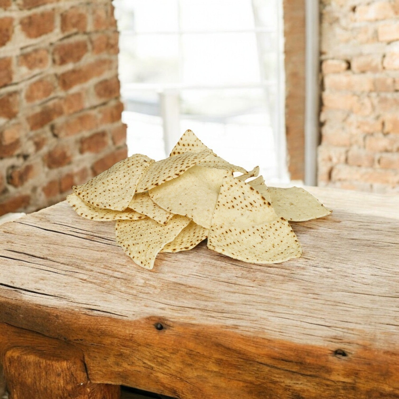 A pile of El Milagro White Corn Tortillas, 4-way cut (1 bag, 20 lbs), sits on a rustic wooden surface with brick walls and a window in the background. Ships on Mondays—please plan accordingly!.